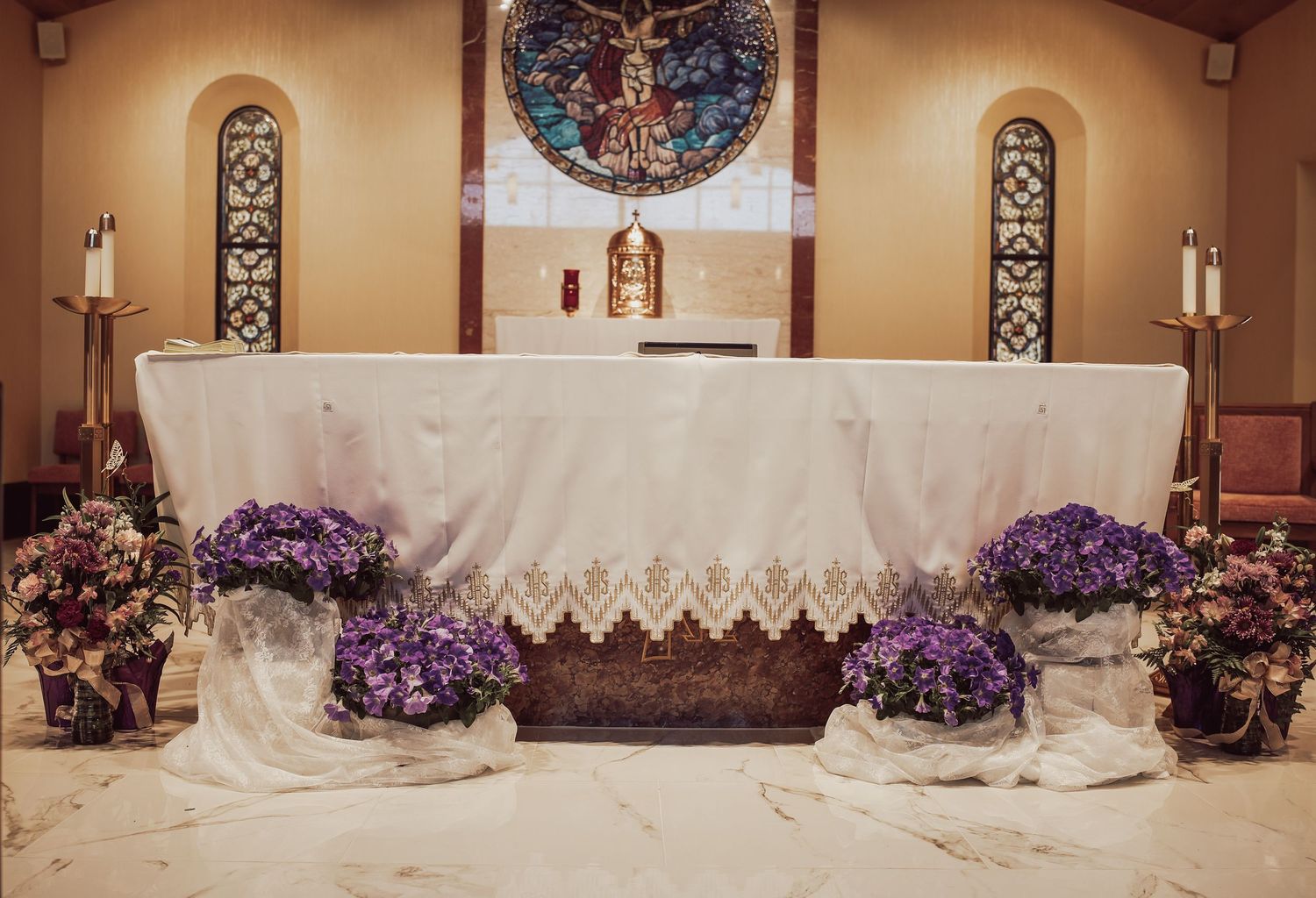 Church altar decorated with white linen, lace trim, and purple floral arrangements in a traditional sanctuary setting.