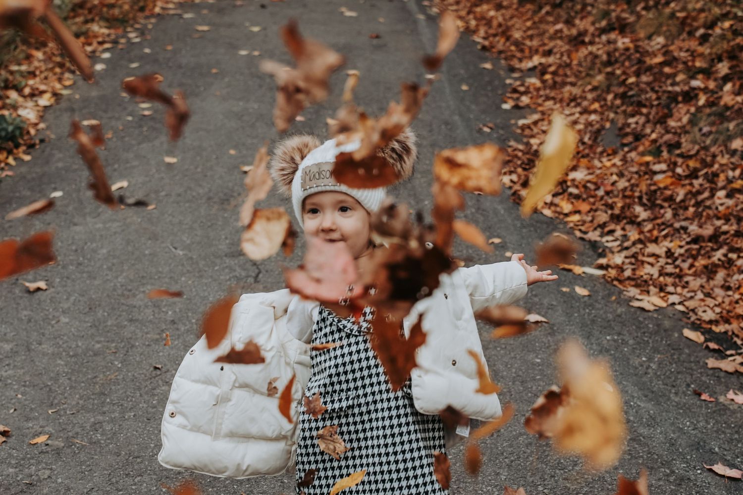 A couple enjoys a playful autumn moment as fallen leaves swirl around them on a path.