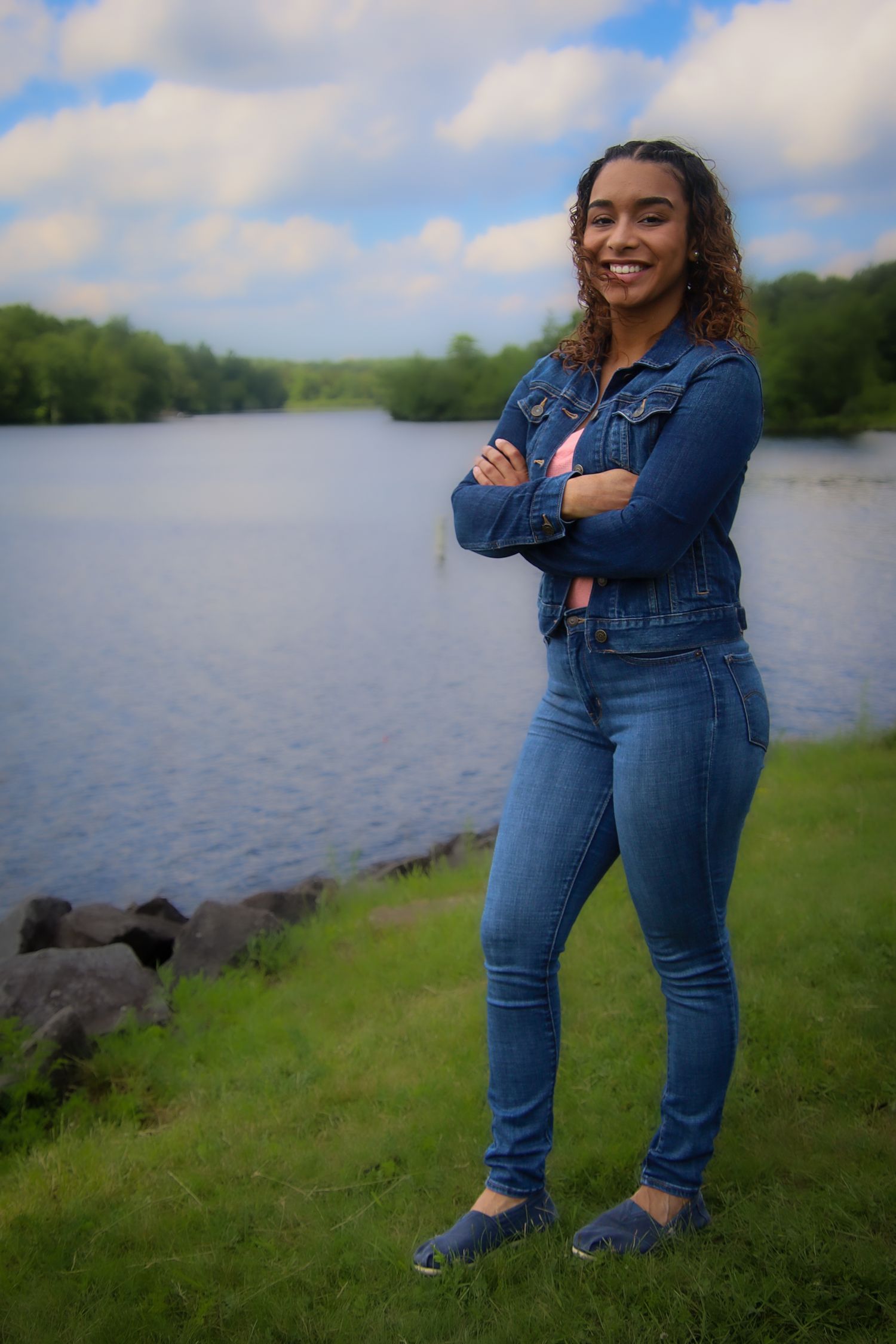 A person in denim clothing poses casually by a scenic lake with green grass and blue sky in the background.