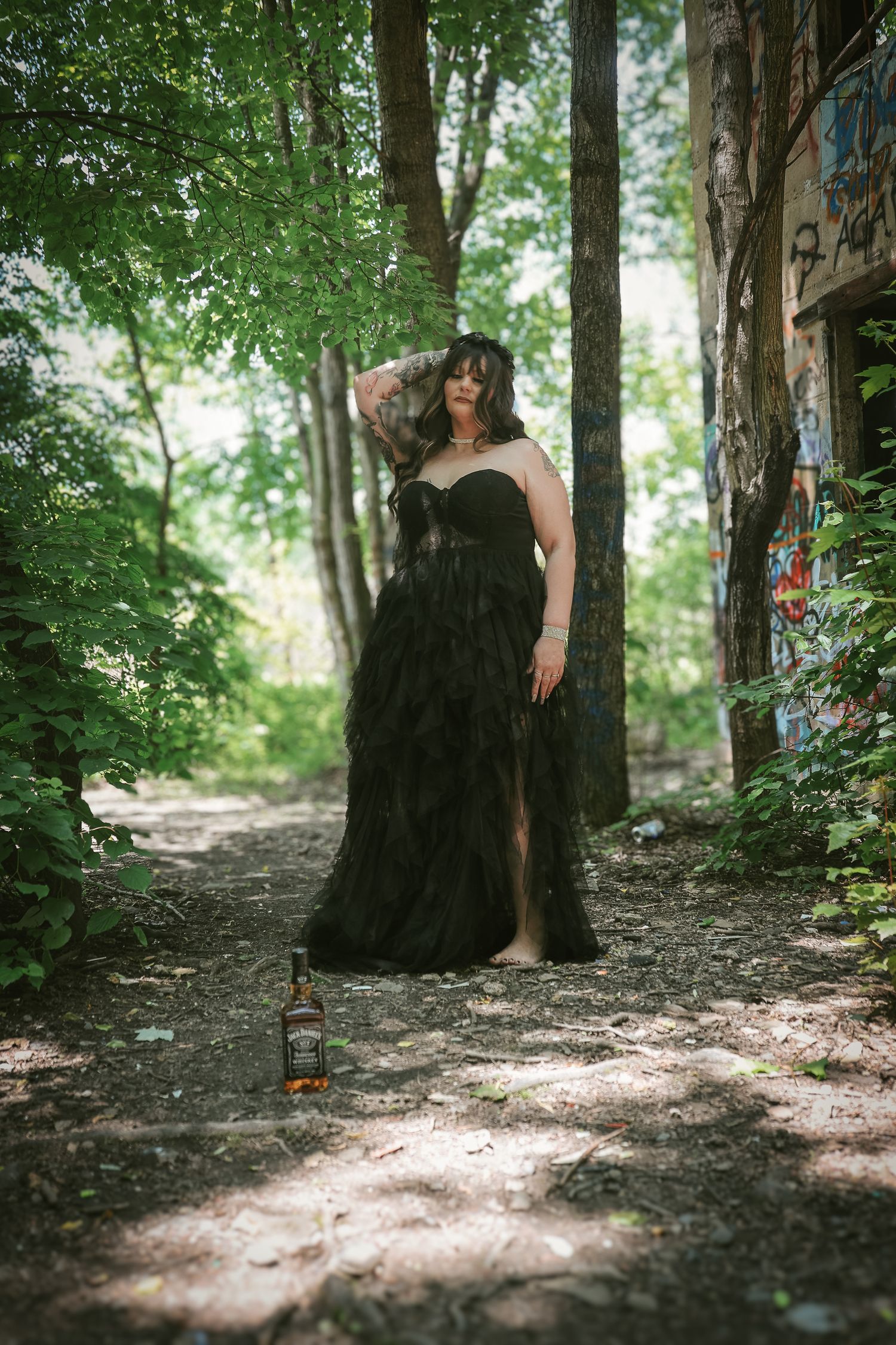 A figure in a flowing black dress stands mysteriously among trees in a wooded forest setting.