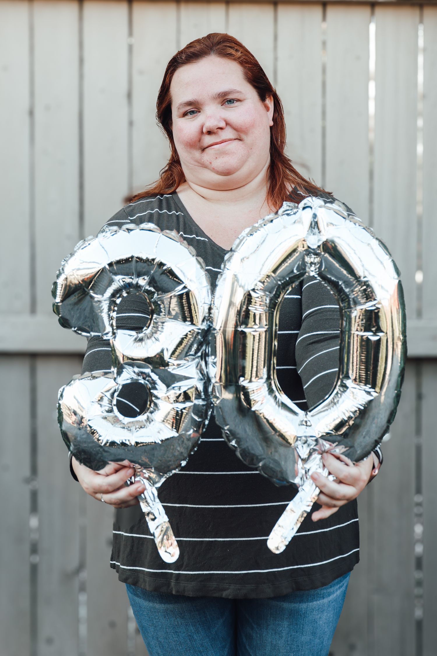 Three silver metallic number 0 balloons being held up against a gray fence background.
