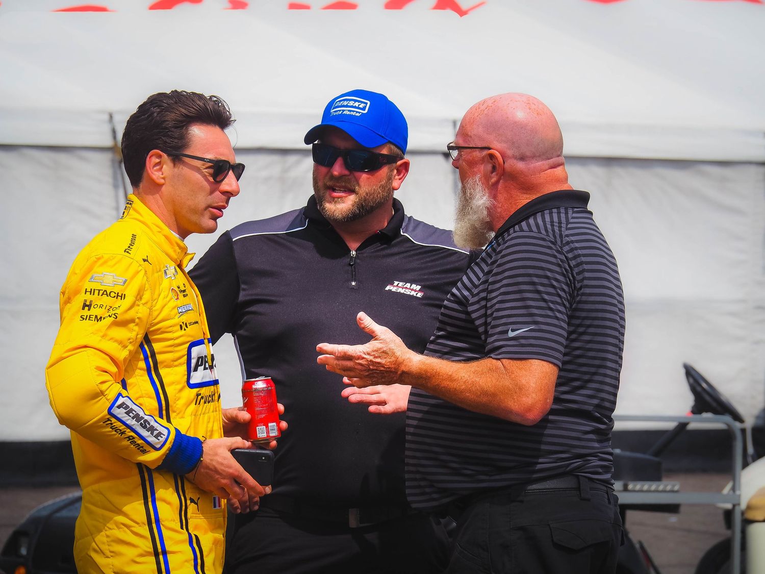 Three people in racing attire having an animated conversation in what appears to be a pit lane or garage area.