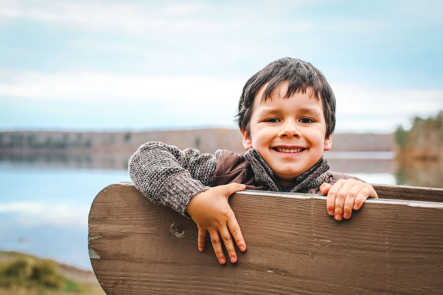 Child wearing a gray sweater leans on wooden railing with lake and cloudy sky in background.