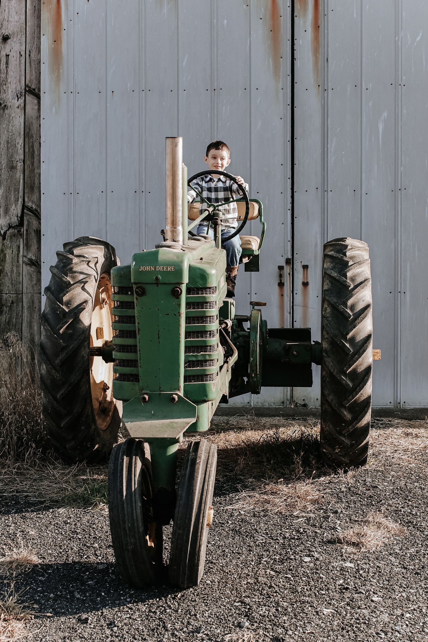 A vintage green tractor parked against a gray wooden wall with gravel ground beneath it.