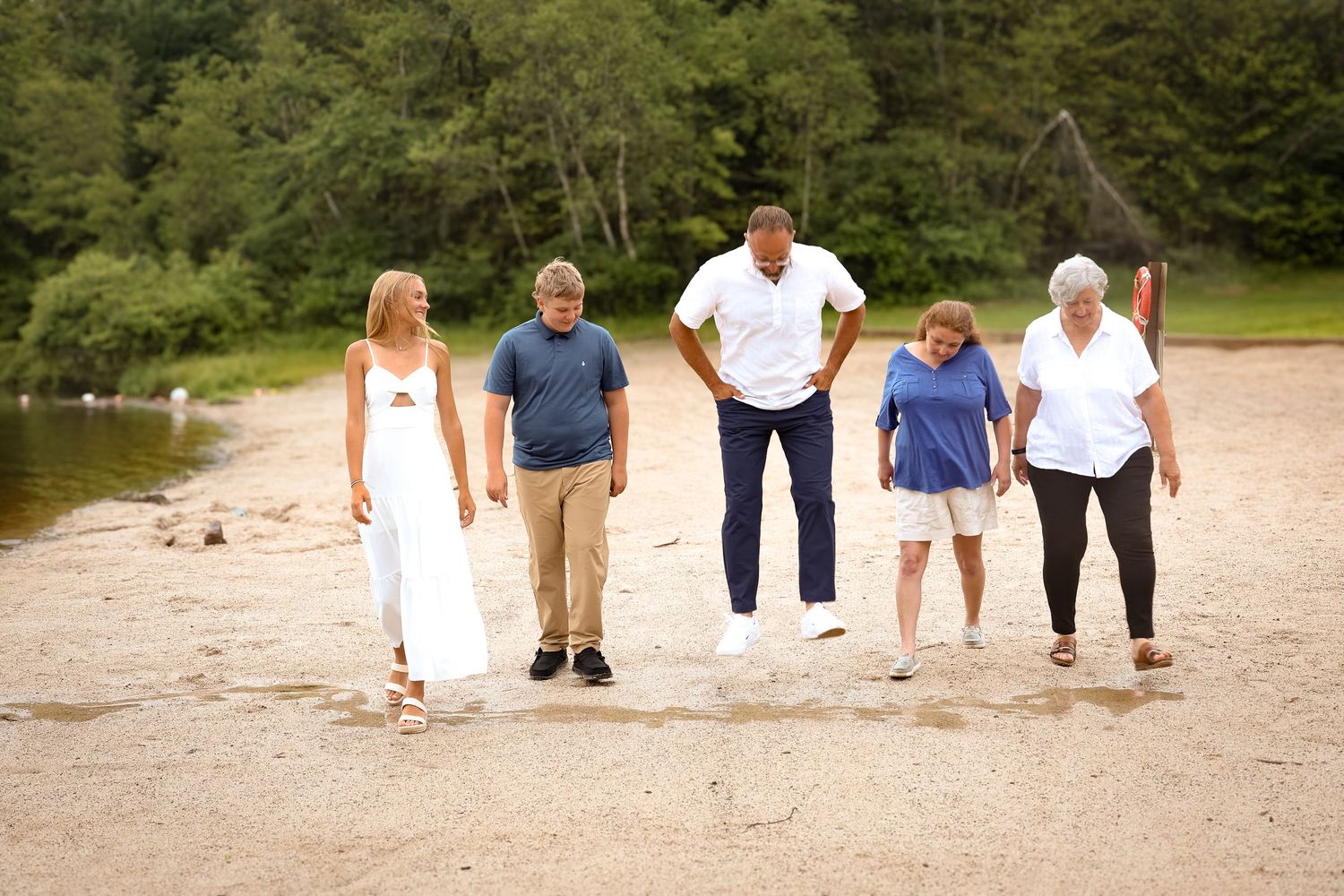 A group of people walking and laughing together on a sandy beach near a wooded shoreline.