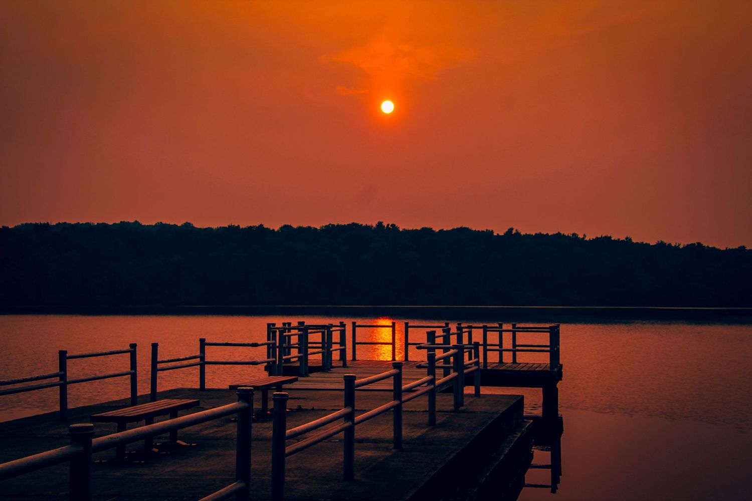 A wooden dock extends into calm waters during an orange sunset with a bright sun visible through smoky skies.