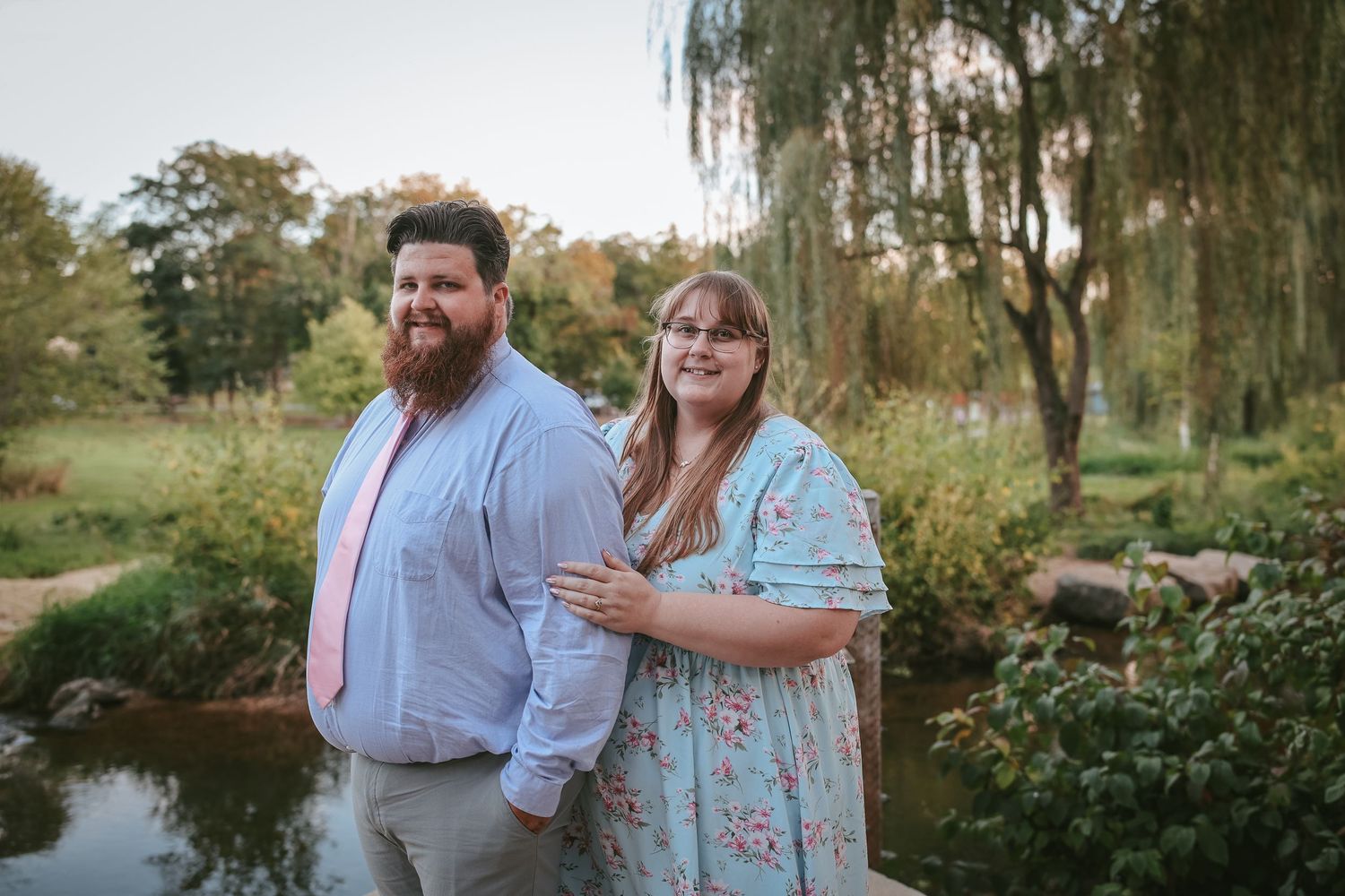 A couple poses together in a romantic outdoor setting with willow trees and a pond in the background.