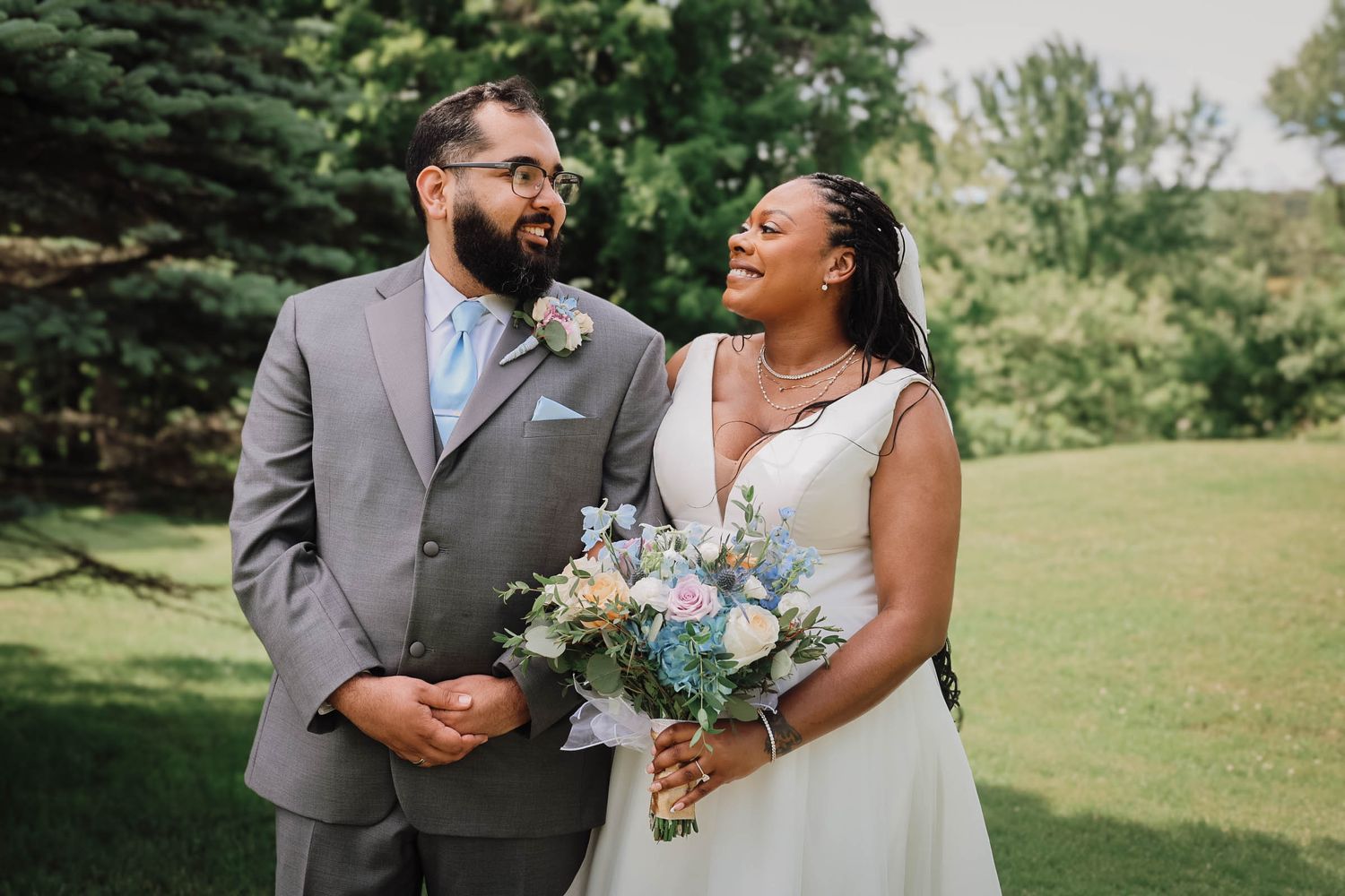 A bride and groom smile joyfully outdoors, she holds a blue and white bouquet, he wears a gray suit with a floral boutonniere.