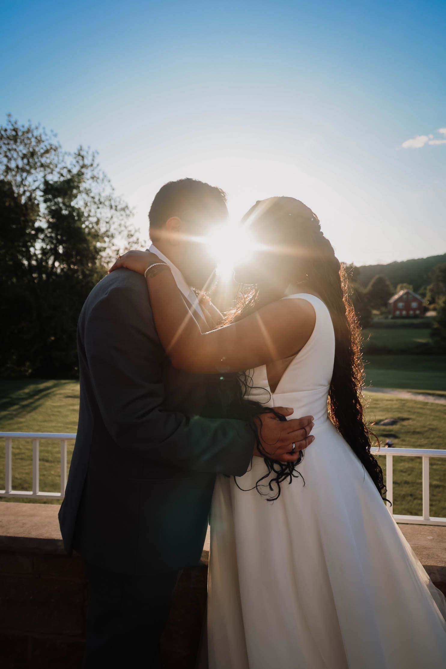 Bride and groom embracing at sunset with golden sunlight glowing between them on a scenic outdoor wedding venue.