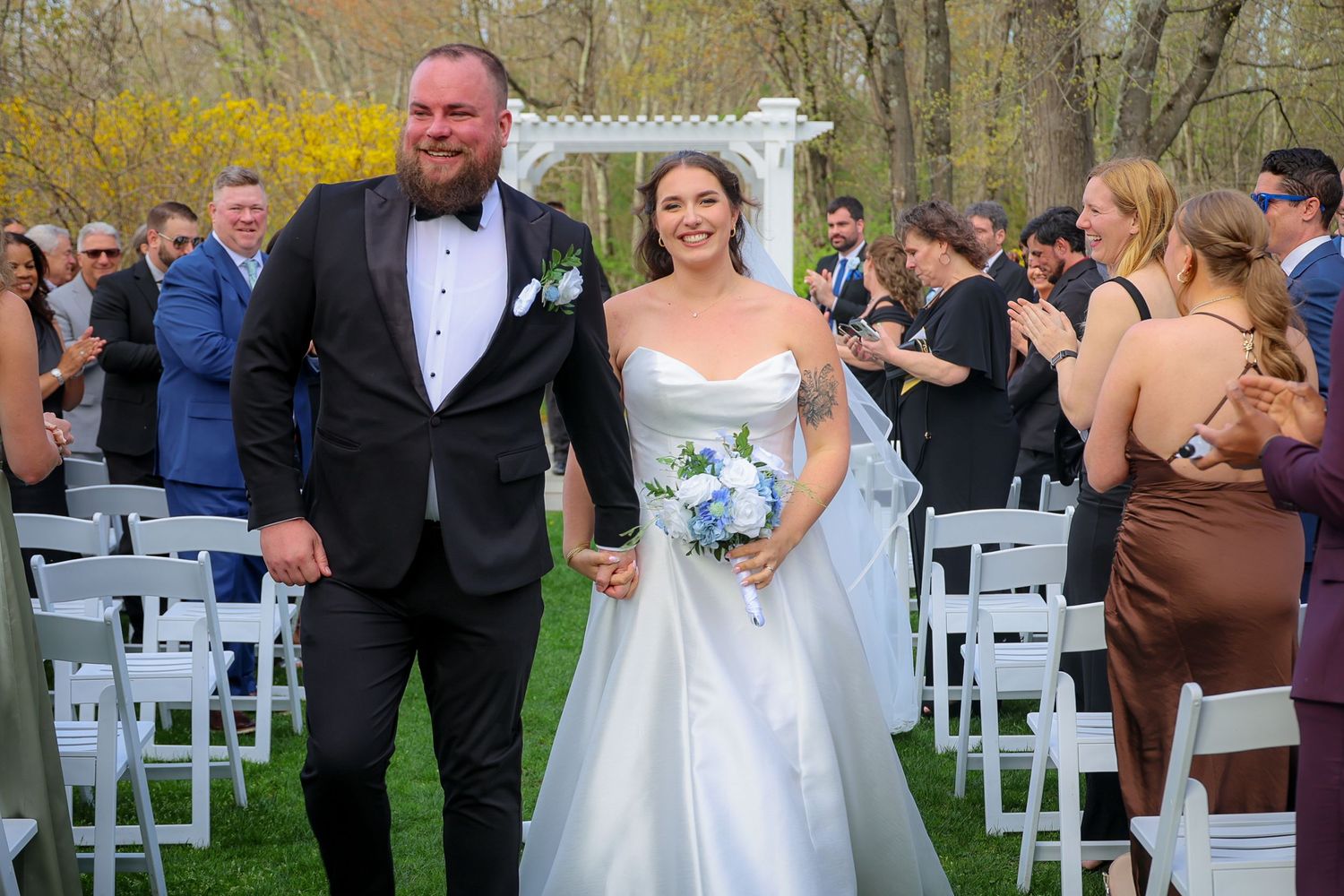 Newlywed couple walks down outdoor wedding aisle, bride in white gown holding bouquet, groom in black tuxedo, guests applauding.