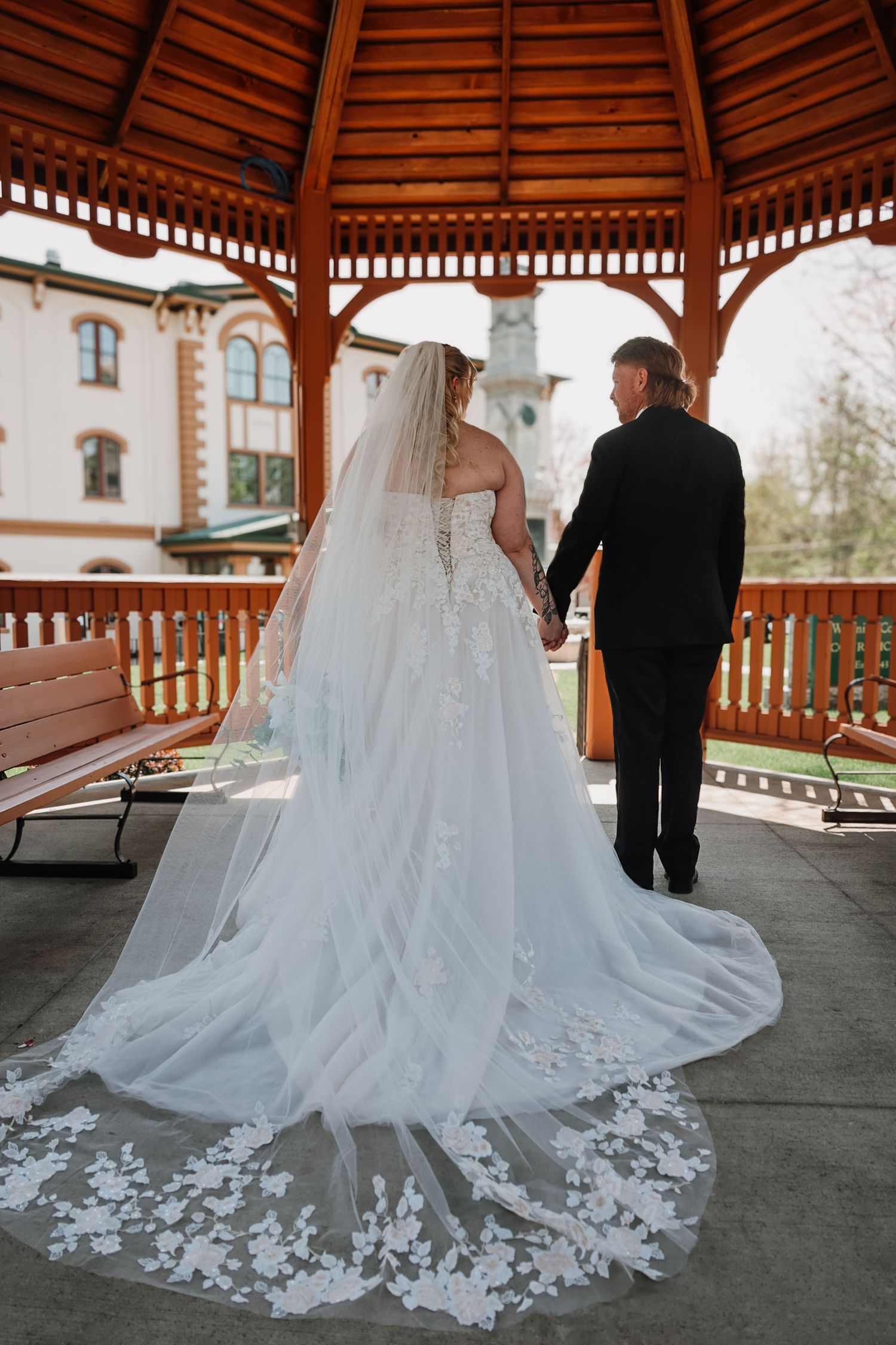 Bride and groom holding hands in a wooden gazebo, showcasing a stunning floral lace cathedral veil and train.