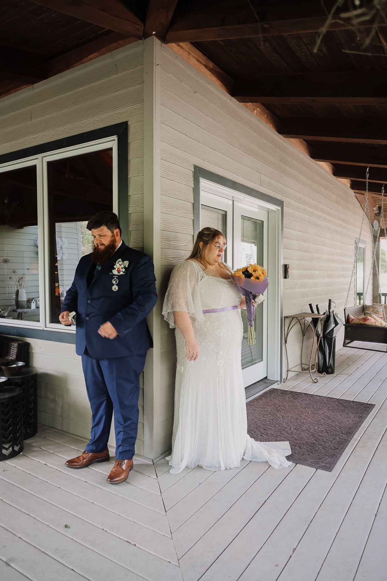 Bride in white gown holding colorful bouquet and groom in navy suit stand on wooden porch before wedding ceremony.