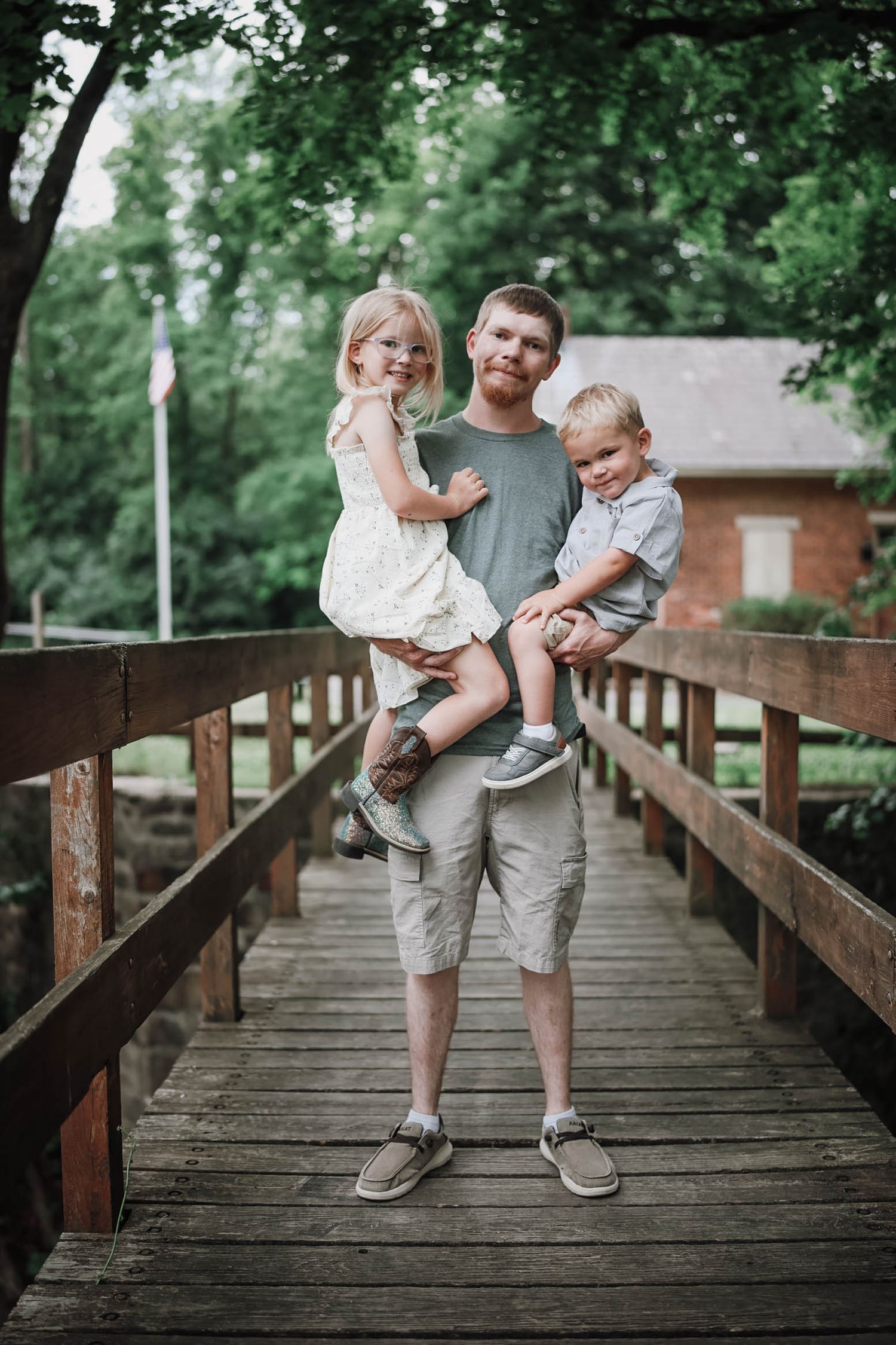 A father holds his two young children on a wooden bridge surrounded by lush green trees near a brick building.