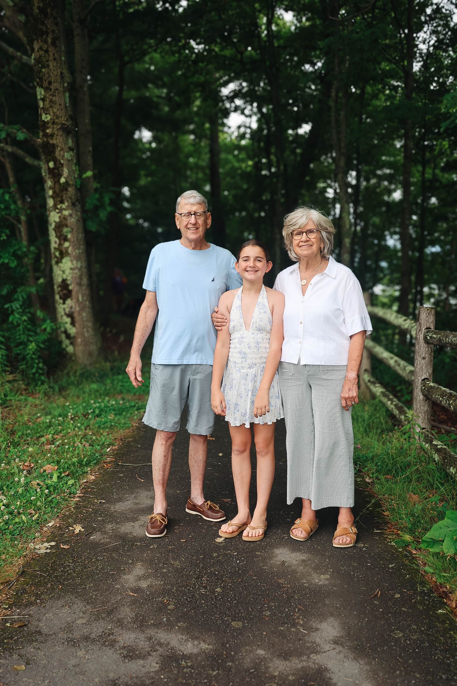 Elderly couple and teenage girl posing together on a tree-lined path outdoors on a summer day.