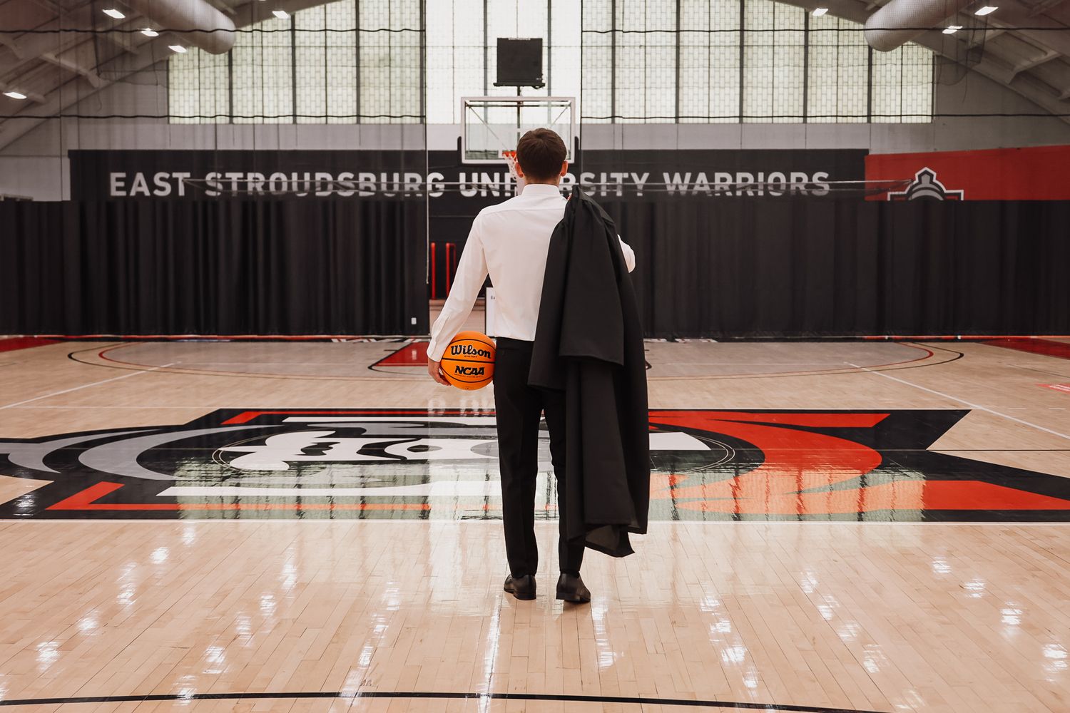 Person holding basketball on East Stroudsburg University Warriors court, viewed from behind in formal attire.