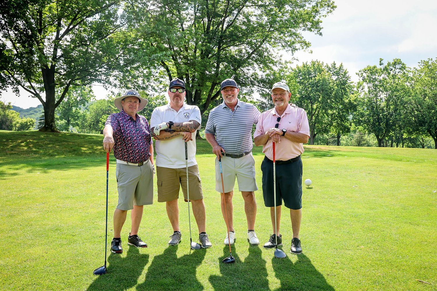 Four men posing together on a lush green golf course on a sunny day, holding golf clubs.