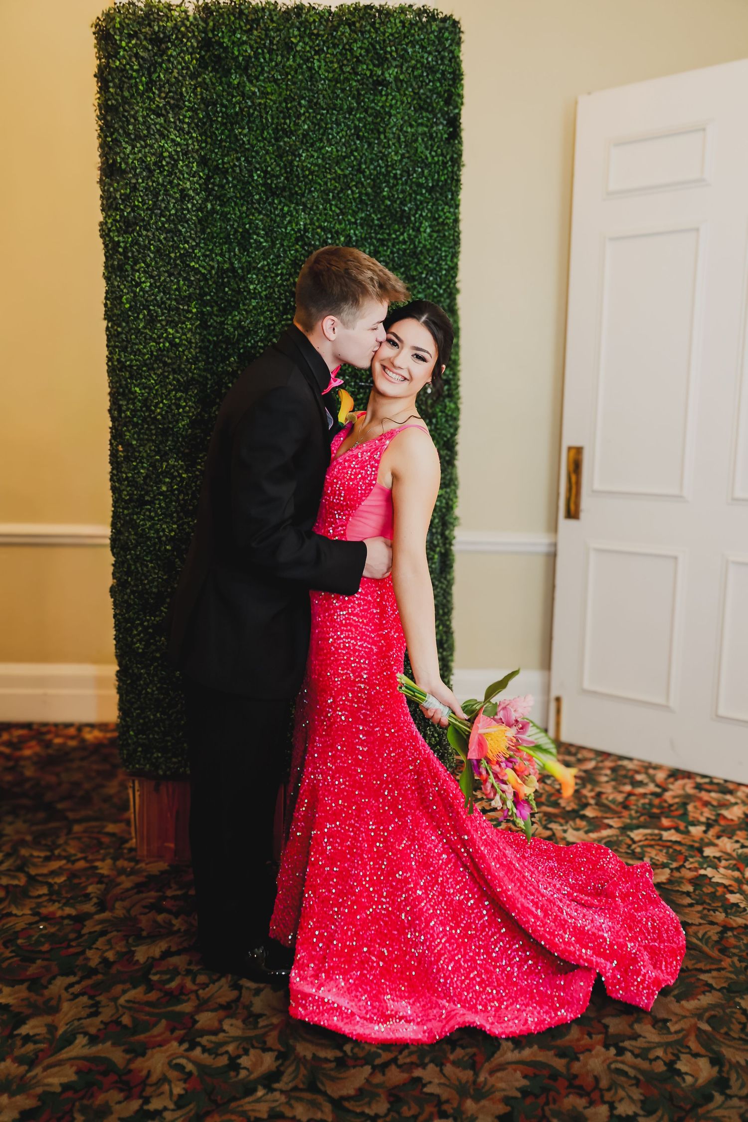 Couple in formal wear, woman in red sequin gown holding bouquet, man in black tuxedo, posing by green hedge wall.