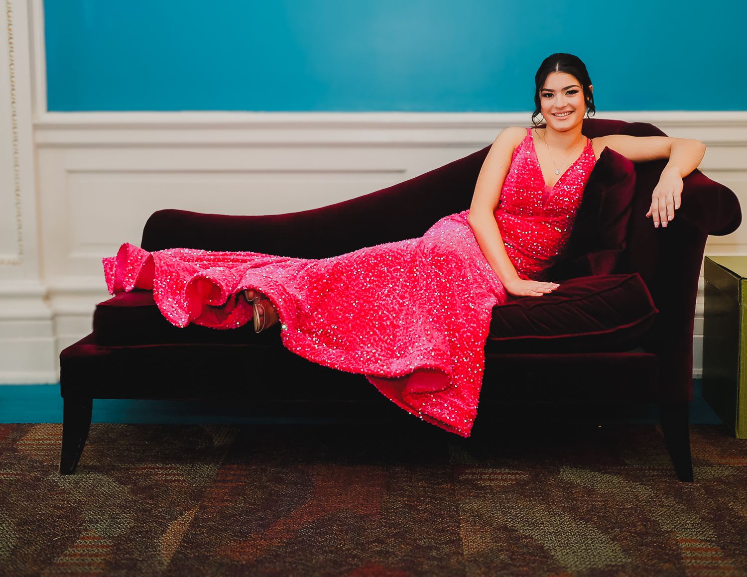 A woman in a stunning pink sequined gown poses elegantly on a dark velvet chaise lounge against a teal wall.