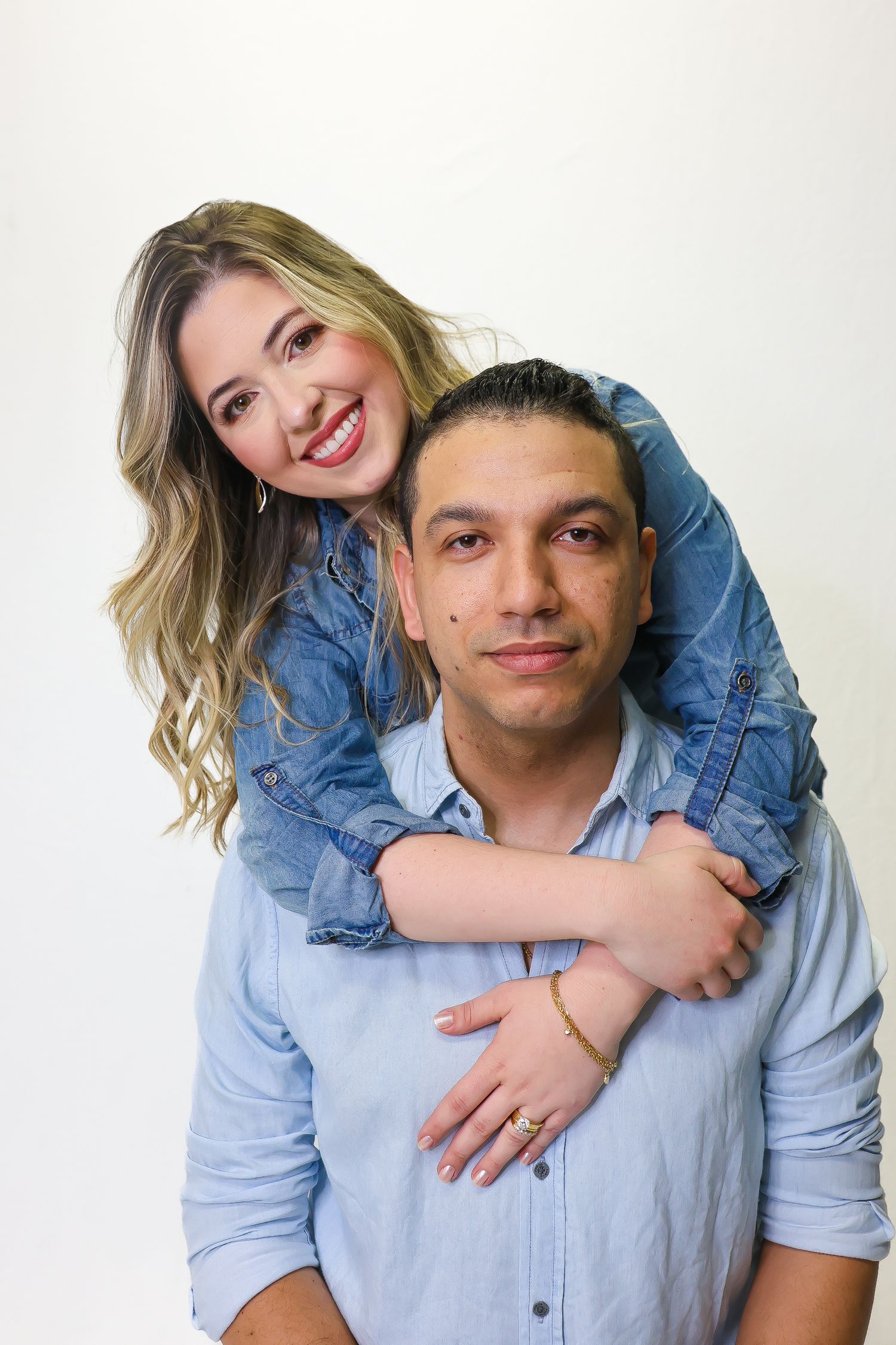 A smiling blonde woman in denim hugs a man from behind against a white background.