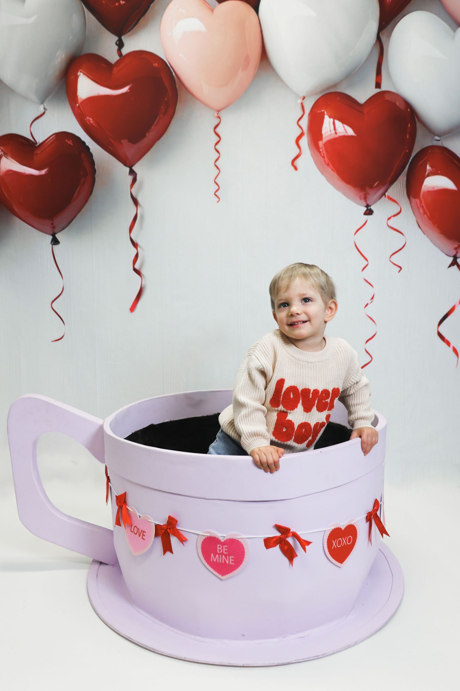 Smiling toddler sits in giant Valentine's Day teacup prop surrounded by red and pink heart-shaped balloons.