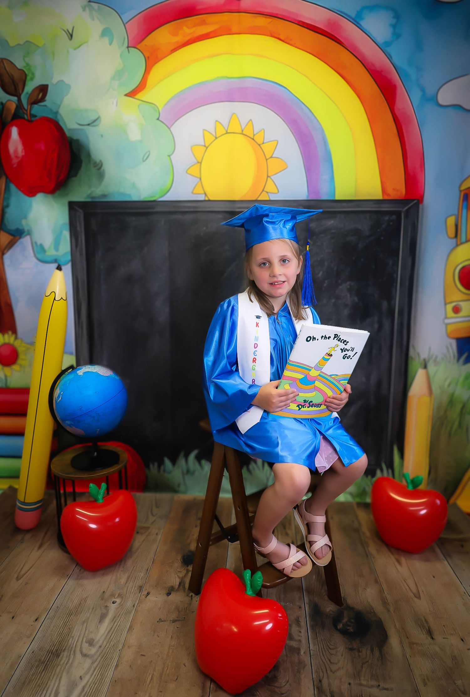 Young girl in blue graduation cap and gown sits on stool holding Dr. Seuss book with colorful school-themed backdrop.