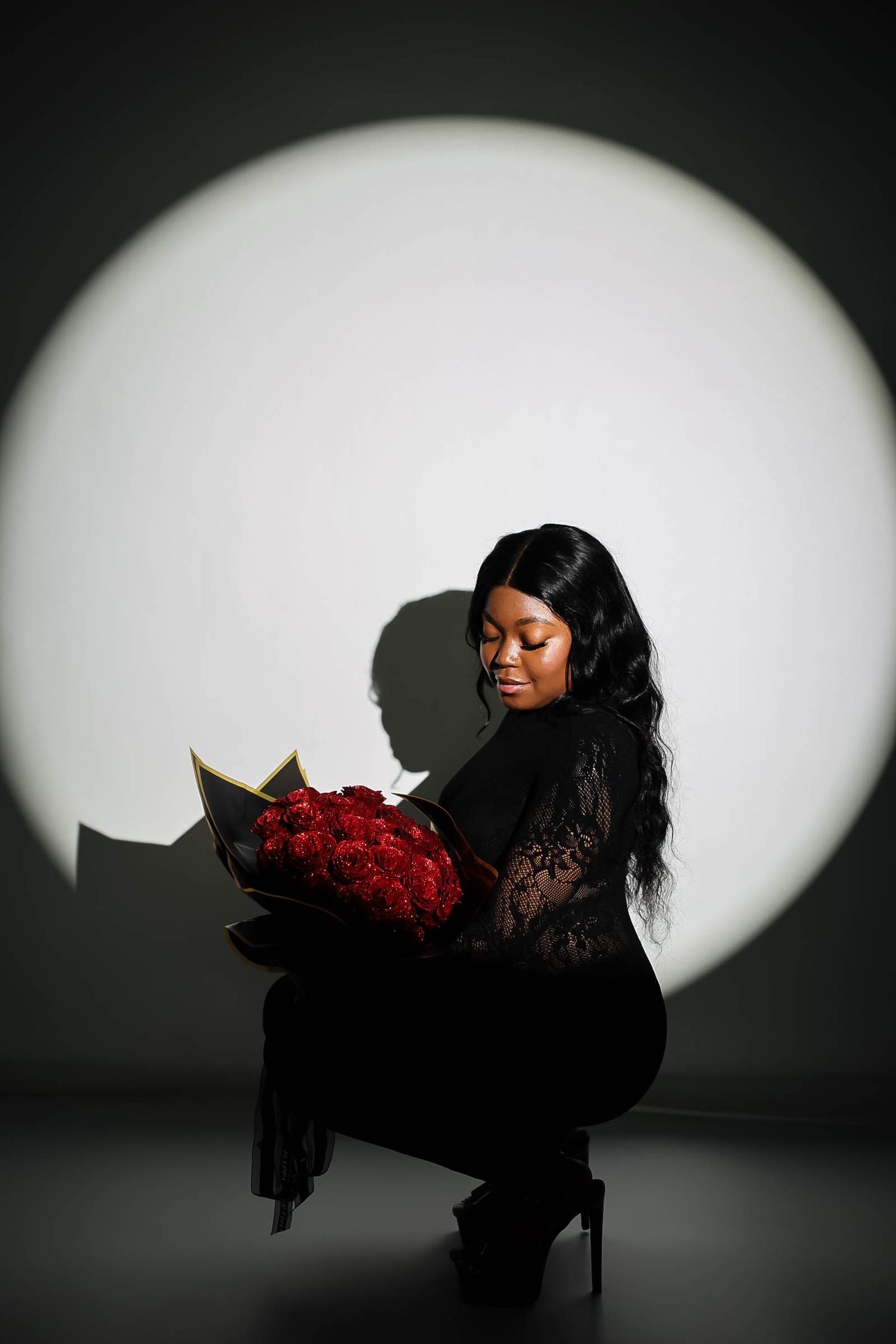Woman in black lace outfit kneeling while admiring red roses bouquet under dramatic spotlight.