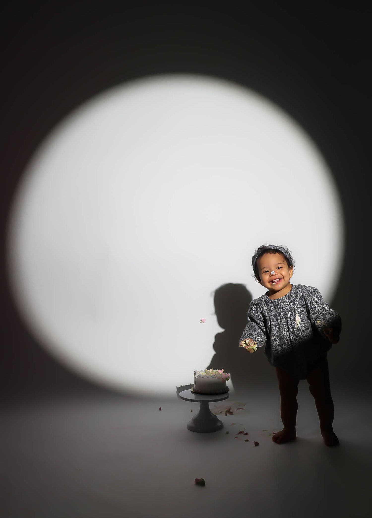 A joyful toddler smiles beside a small cake in a dramatic spotlight, celebrating their first birthday.