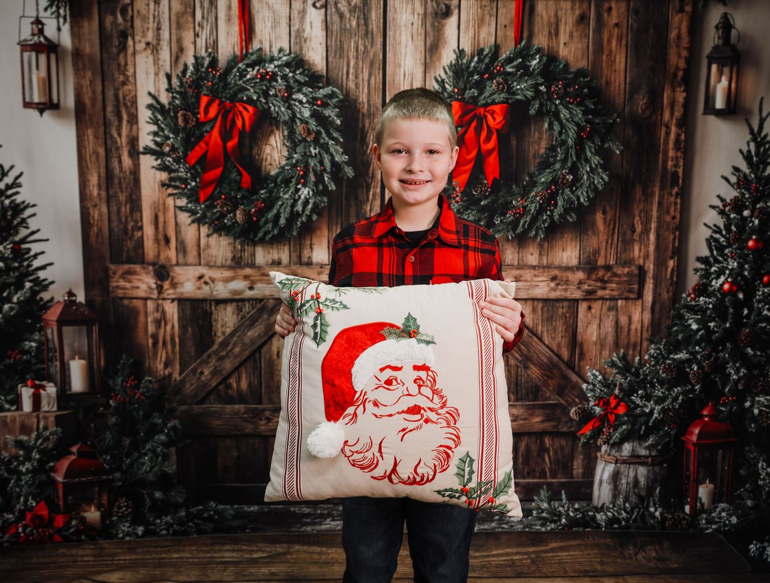 A smiling boy in a red plaid shirt holds a Santa Claus pillow in front of a rustic Christmas barn door backdrop.
