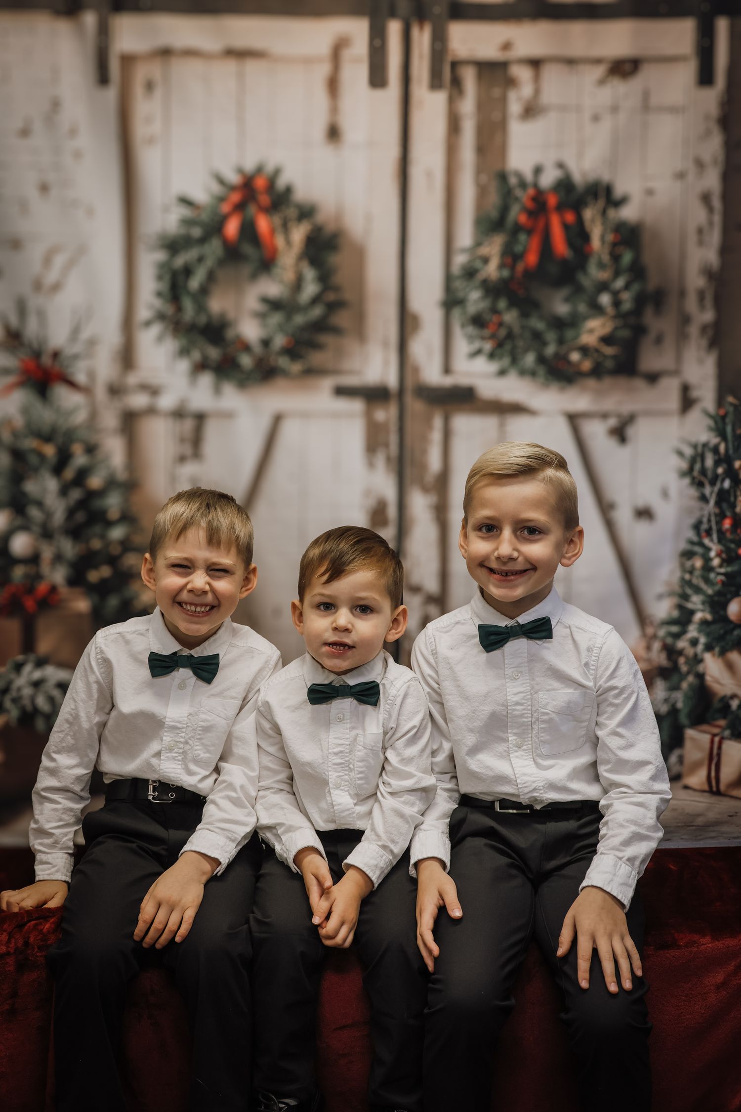Three boys in white shirts and green bow ties pose for a festive Christmas photo in front of holiday wreaths and decorations.