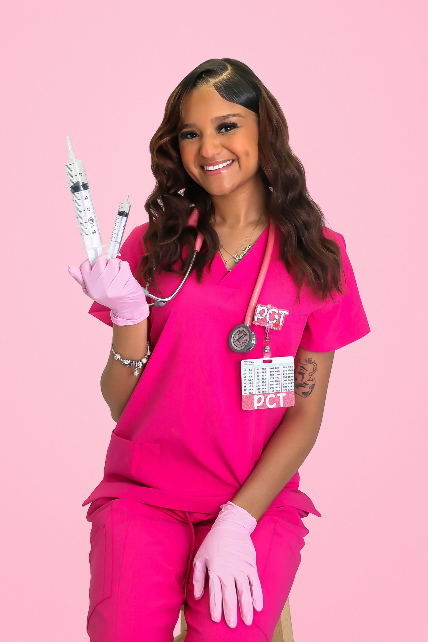 Smiling nurse in pink scrubs holding syringes with stethoscope and DCT badge against pink background.
