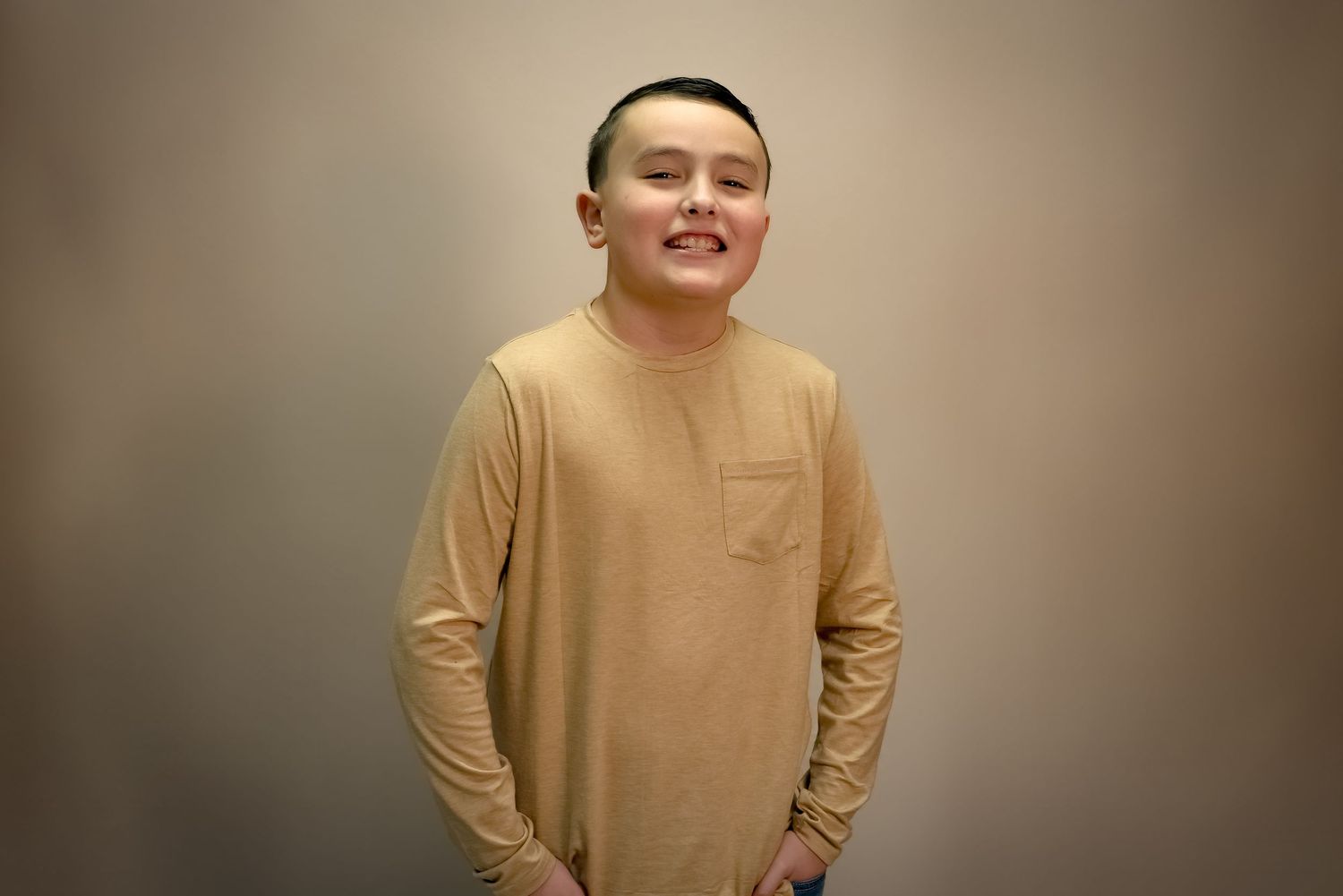 Young Asian man smiling in a beige long-sleeve shirt, standing against a neutral gray background.