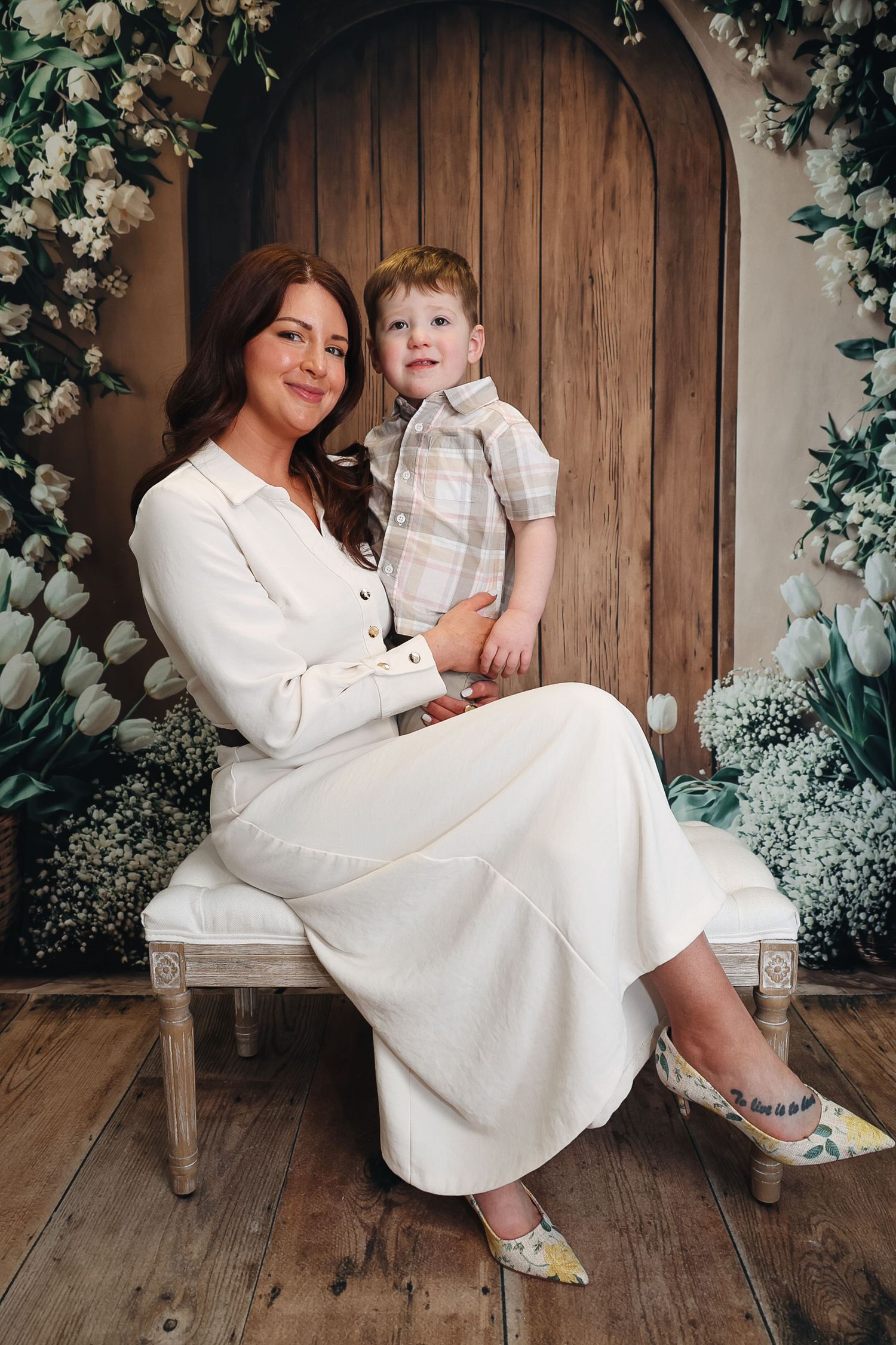 A woman in white dress sits with a young red-haired boy among white tulips and floral backdrop.