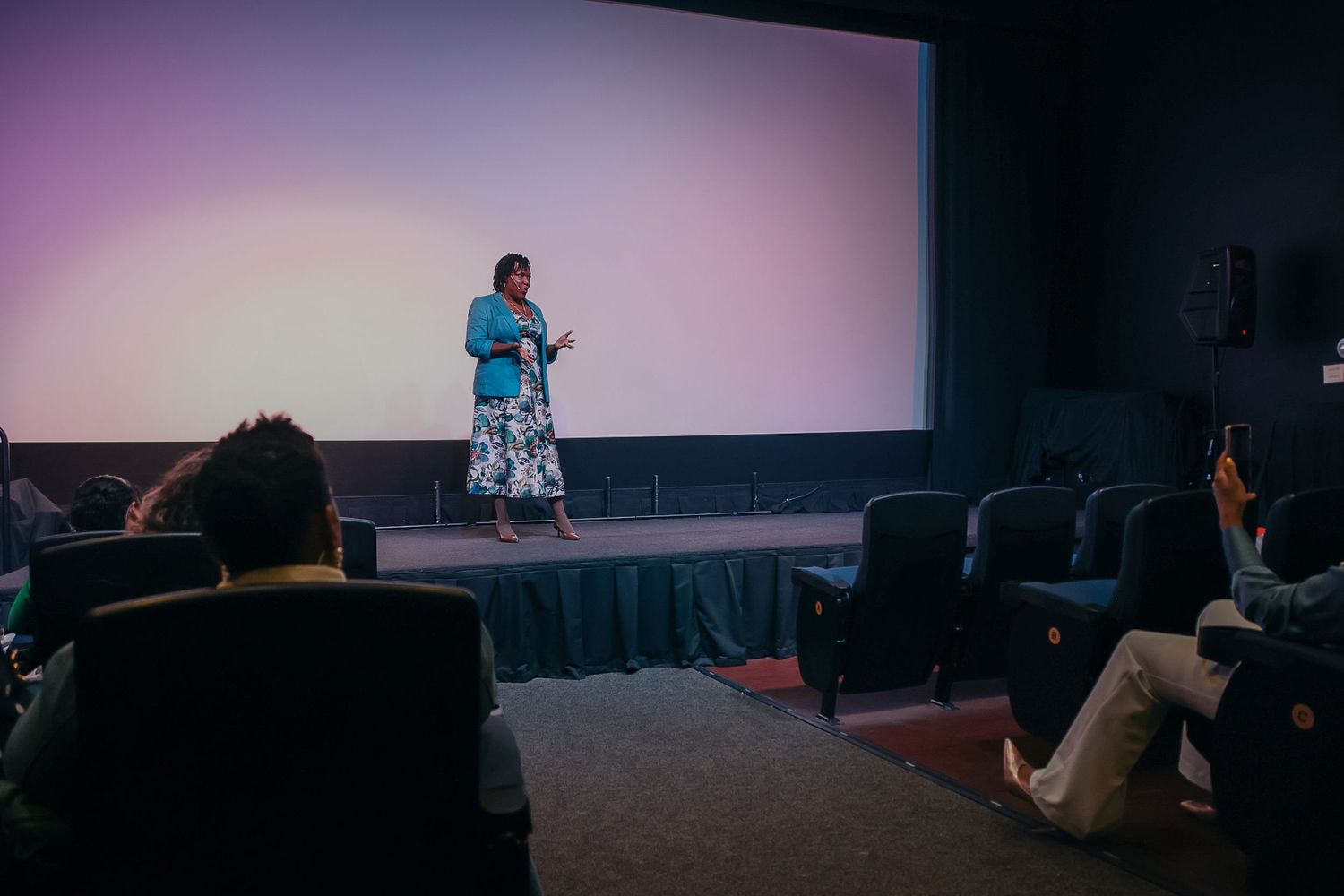 Woman in blue top presents before a blank screen in a small cinema with seated audience members.