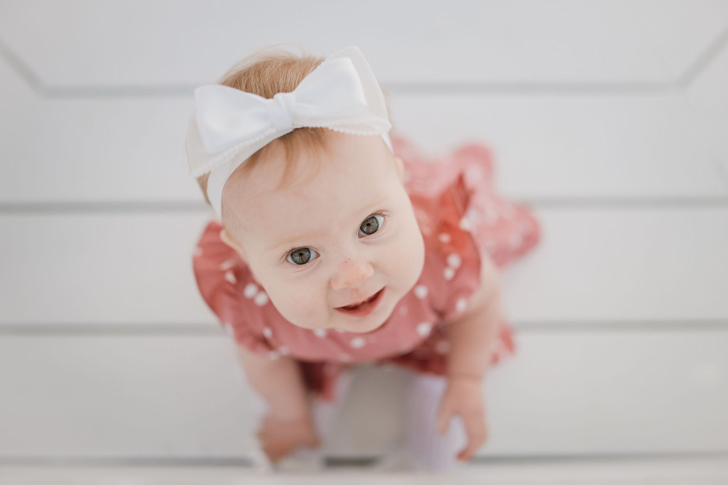 Adorable baby girl in pink polka dot dress and white bow headband smiling up at camera on white background.