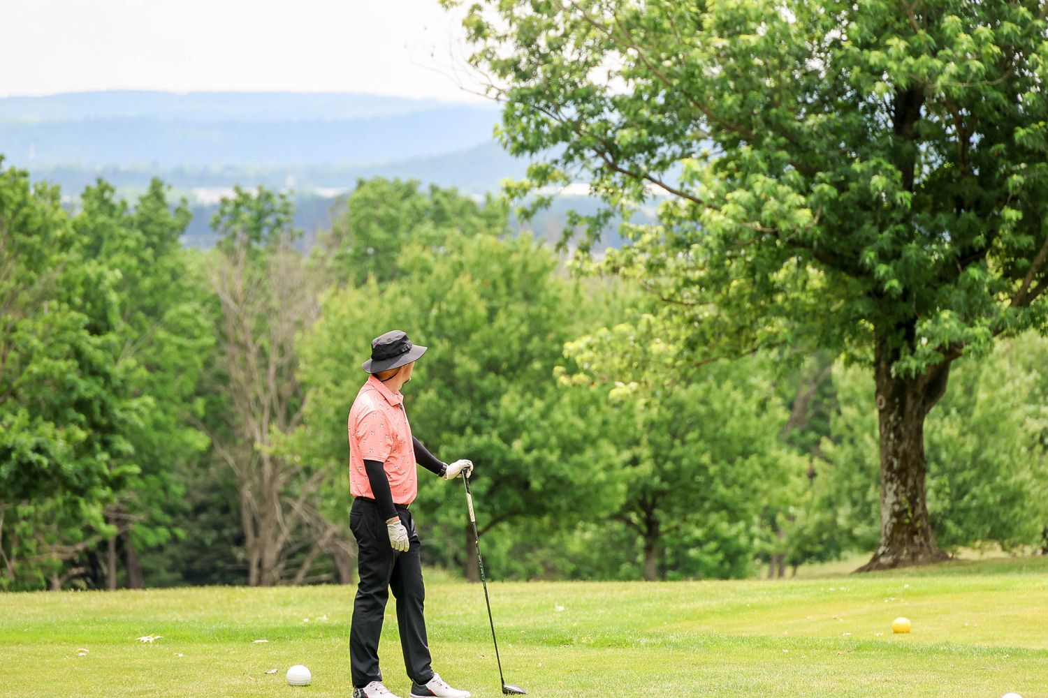 Golfer in pink shirt standing on lush green golf course with scenic tree-lined hills backdrop.