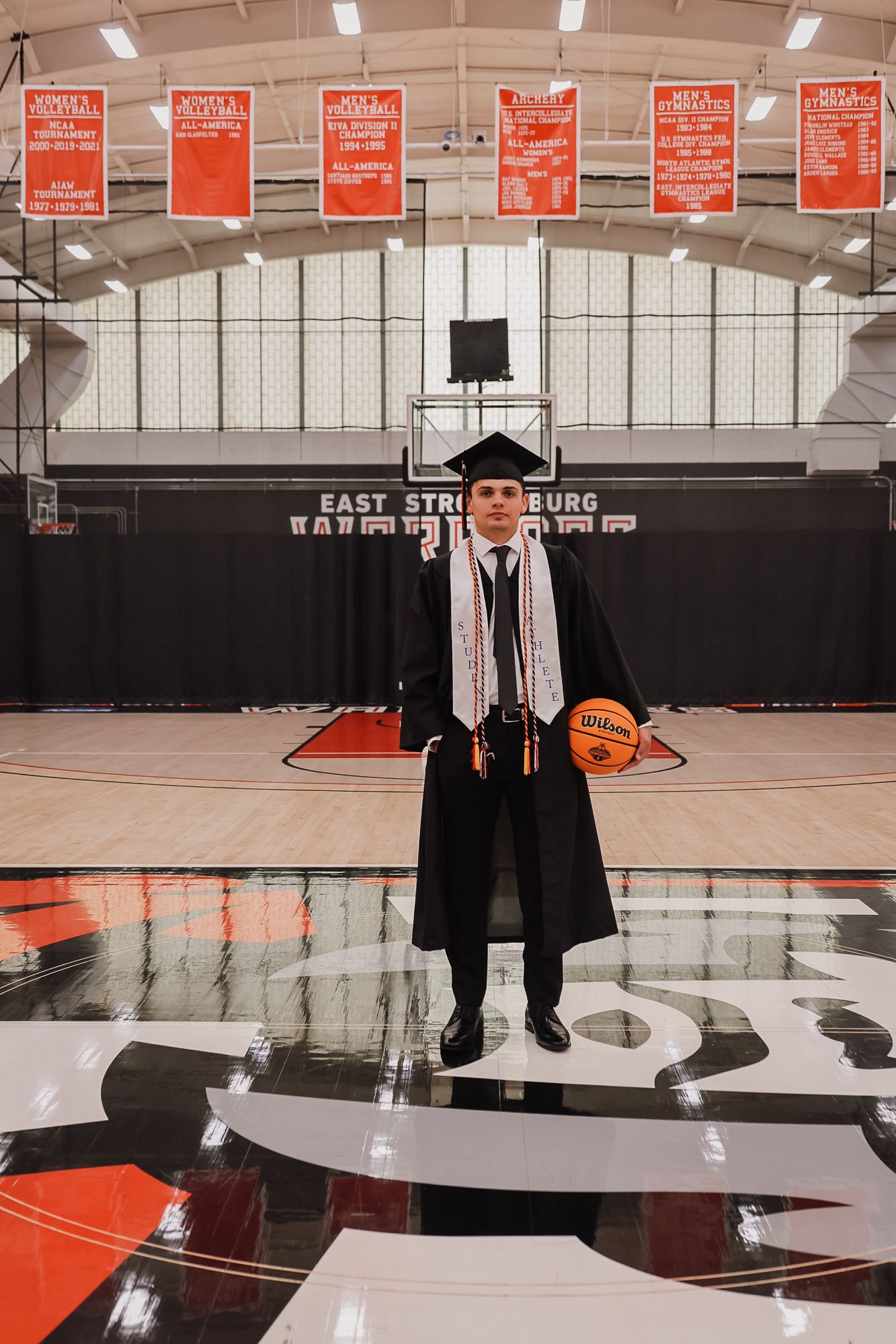 Graduate in cap and gown holds basketball on East Stroudsburg University court, celebrating academic and athletic achievement.