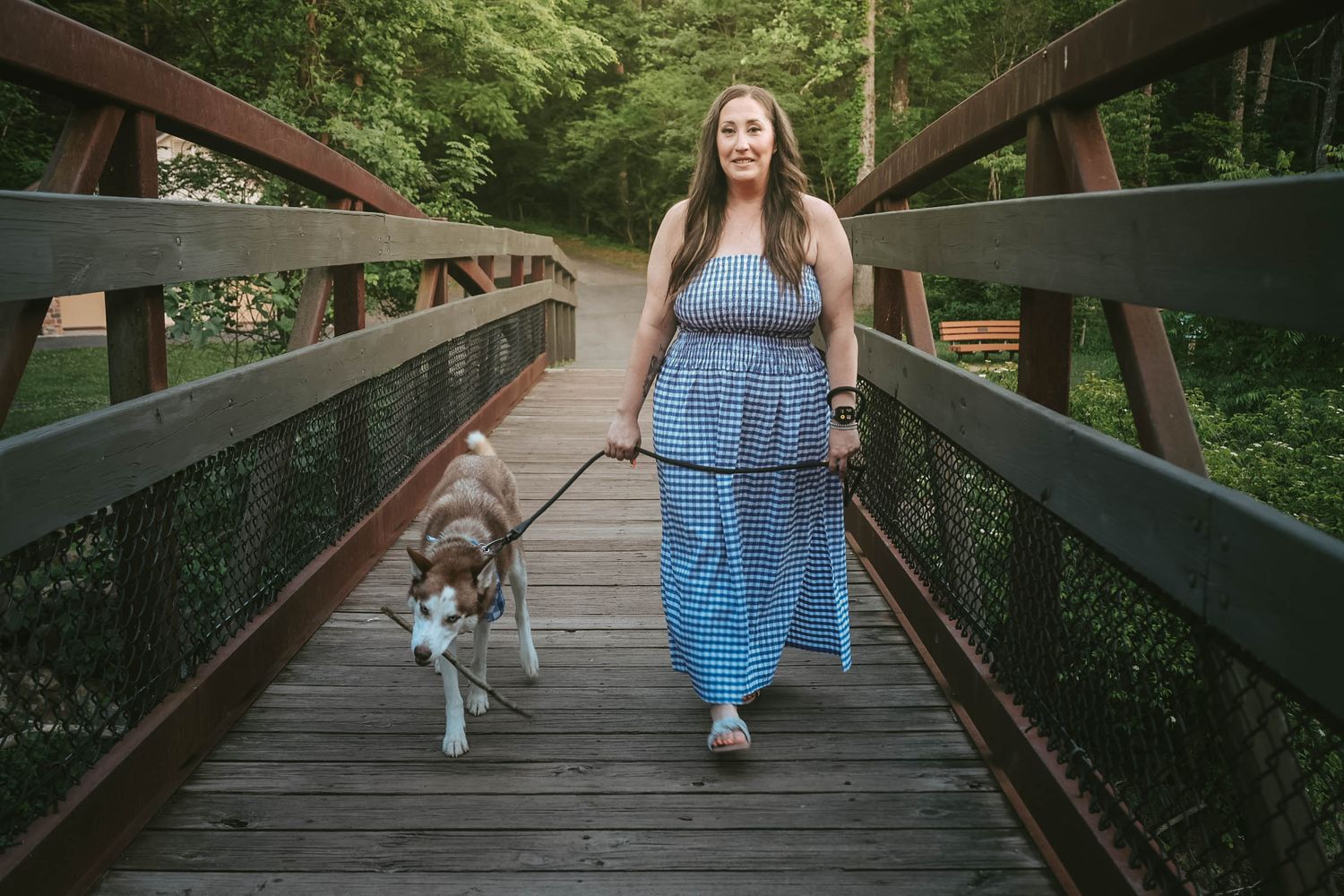 Woman in blue maxi dress walking a dog on a wooden bridge surrounded by lush green trees.