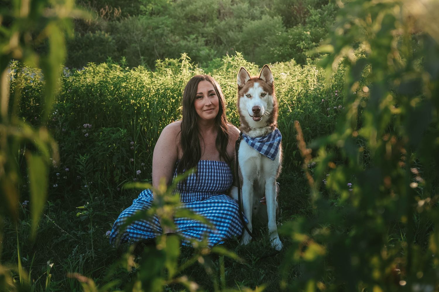Woman in blue gingham dress sitting with Husky dog wearing bandana in lush green field.