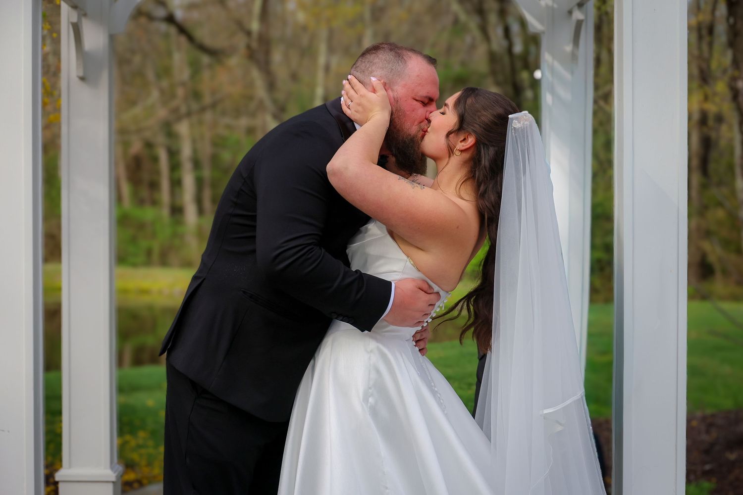 Bride and groom share their first kiss under a white gazebo arch, surrounded by lush green trees on their wedding day.