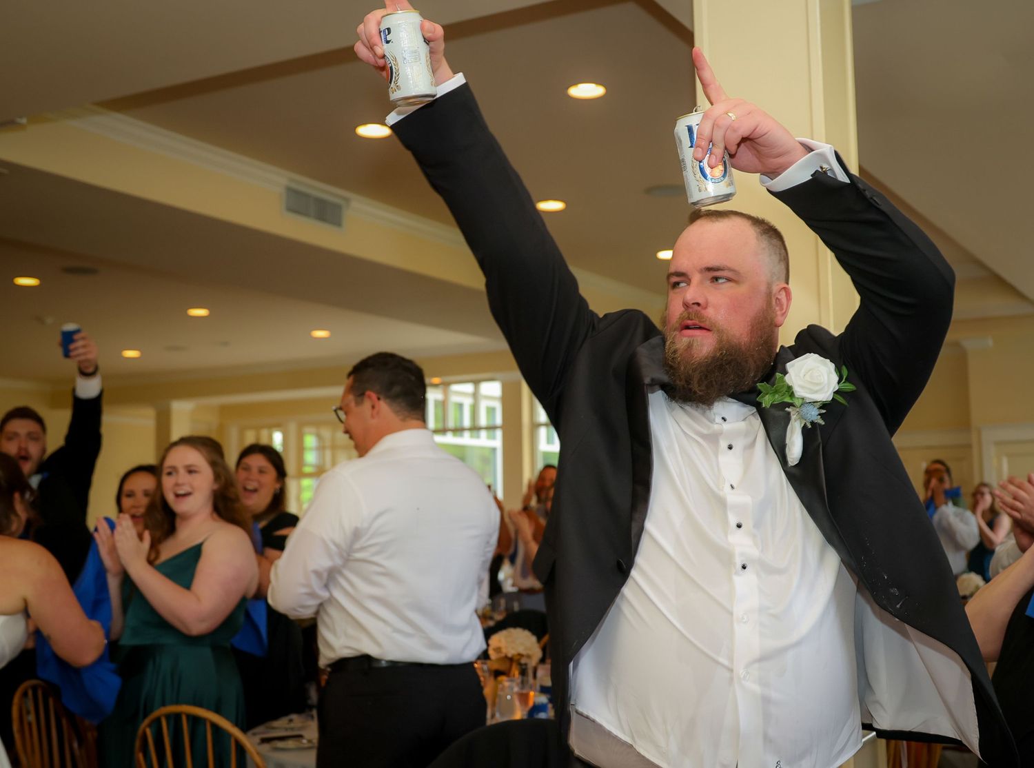 Bearded groom raises arms in celebration at wedding reception, holding drink, wearing tuxedo with white boutonniere.