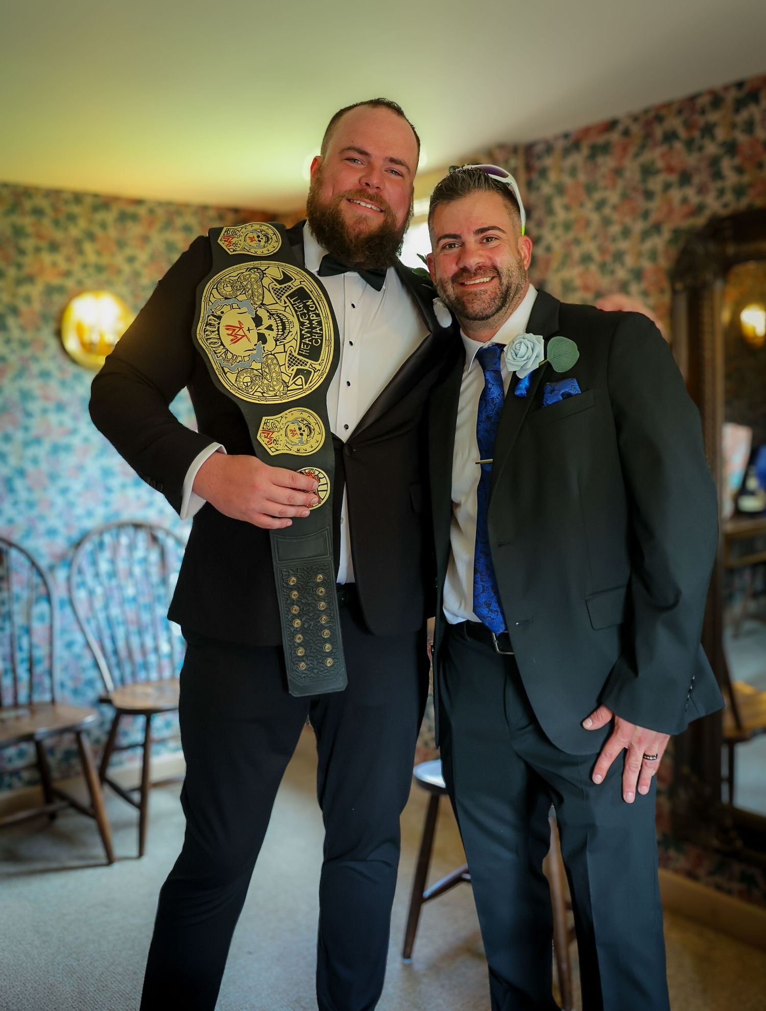Two men in suits pose with a championship wrestling belt at a formal event.