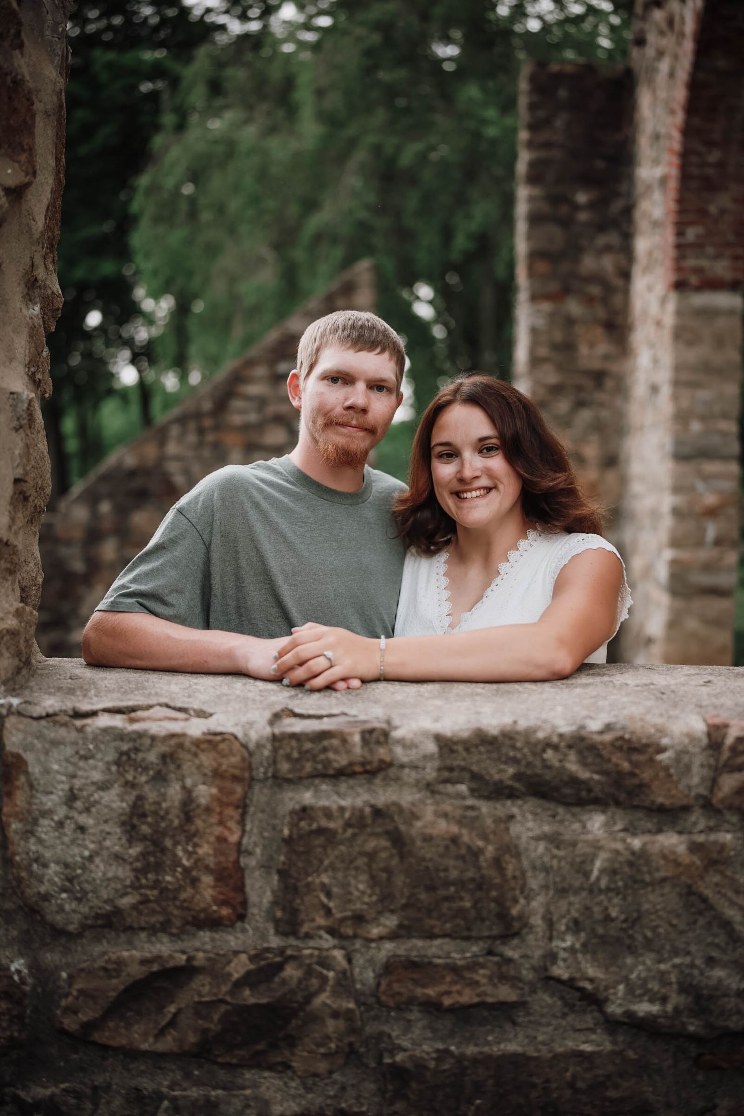 Smiling couple leaning on a rustic stone wall outdoors, surrounded by lush green trees.