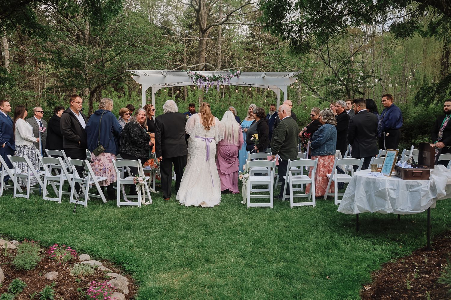 Outdoor wedding ceremony on lush green lawn with white chairs, floral arbor, bride and groom, and guests gathered in garden setting.