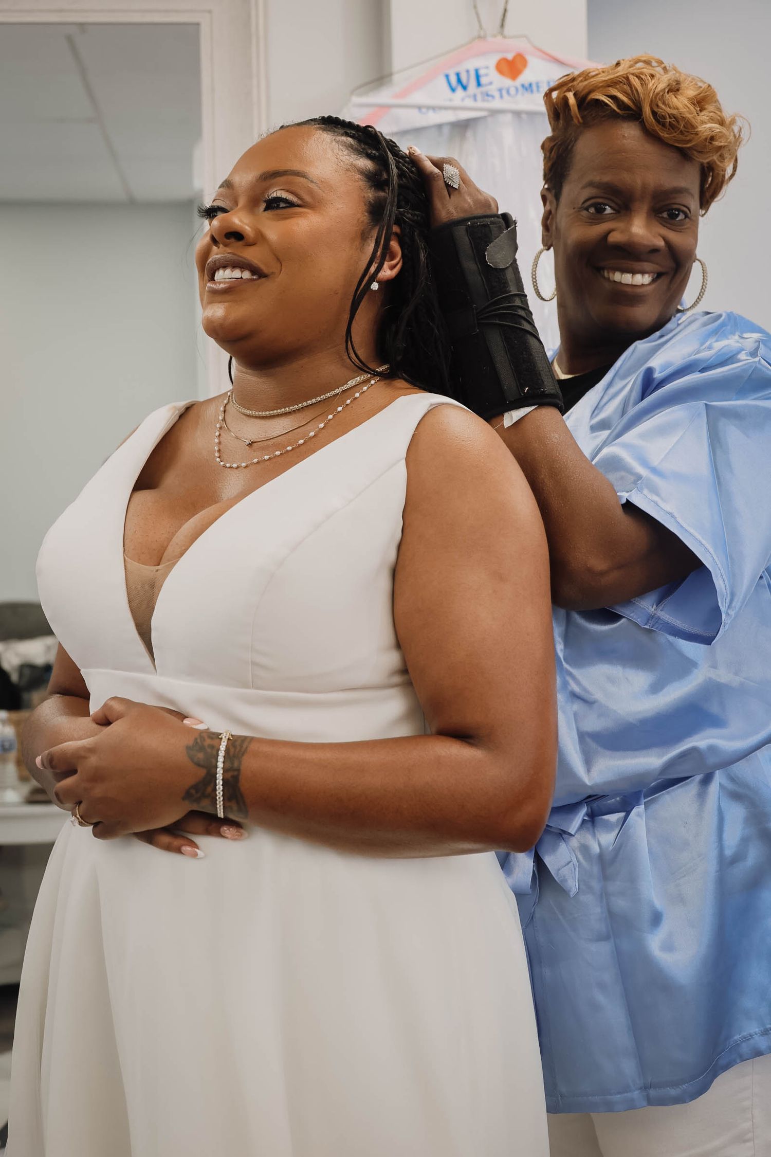 Smiling bride in white dress gets her hair styled by a woman in blue during wedding day preparations.