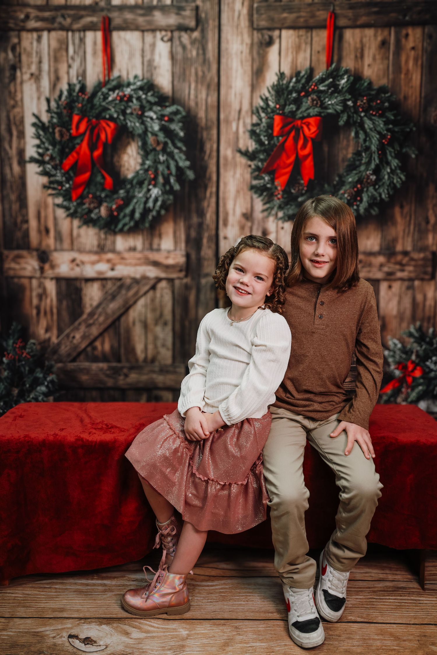Two children pose for a Christmas photo in front of rustic wooden doors adorned with festive wreaths and red bows.