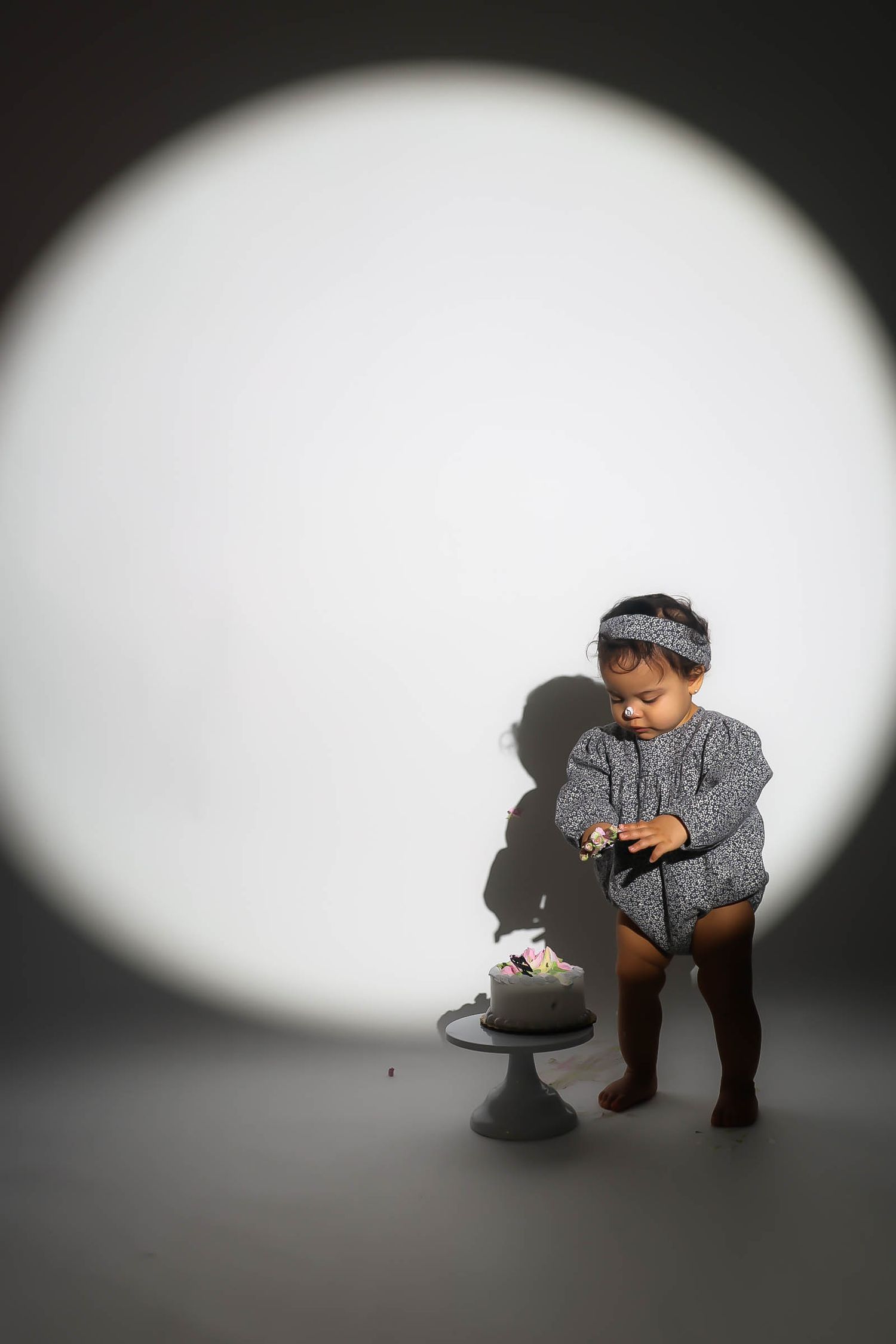 Toddler girl in gray outfit with headband stands beside a small cake on a pedestal, illuminated by a circular spotlight.