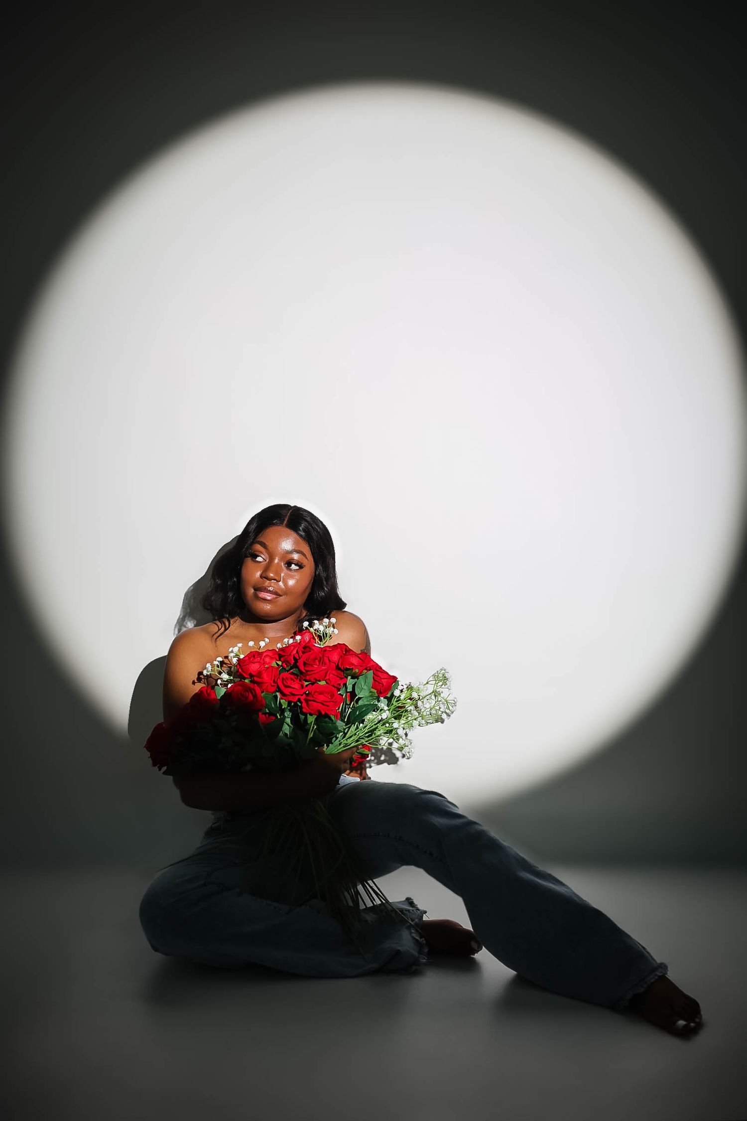 Woman sitting on floor holding large red roses bouquet, wearing jeans, posed against white circular spotlight background.