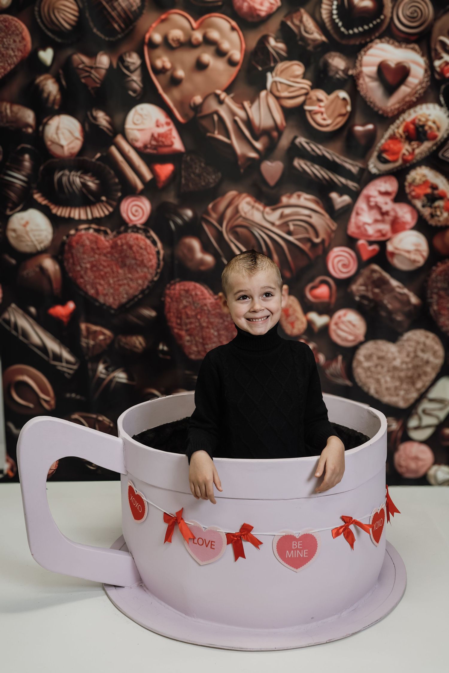 Smiling child sitting in a giant teacup prop with Valentine's Day decorations and chocolate candy backdrop.