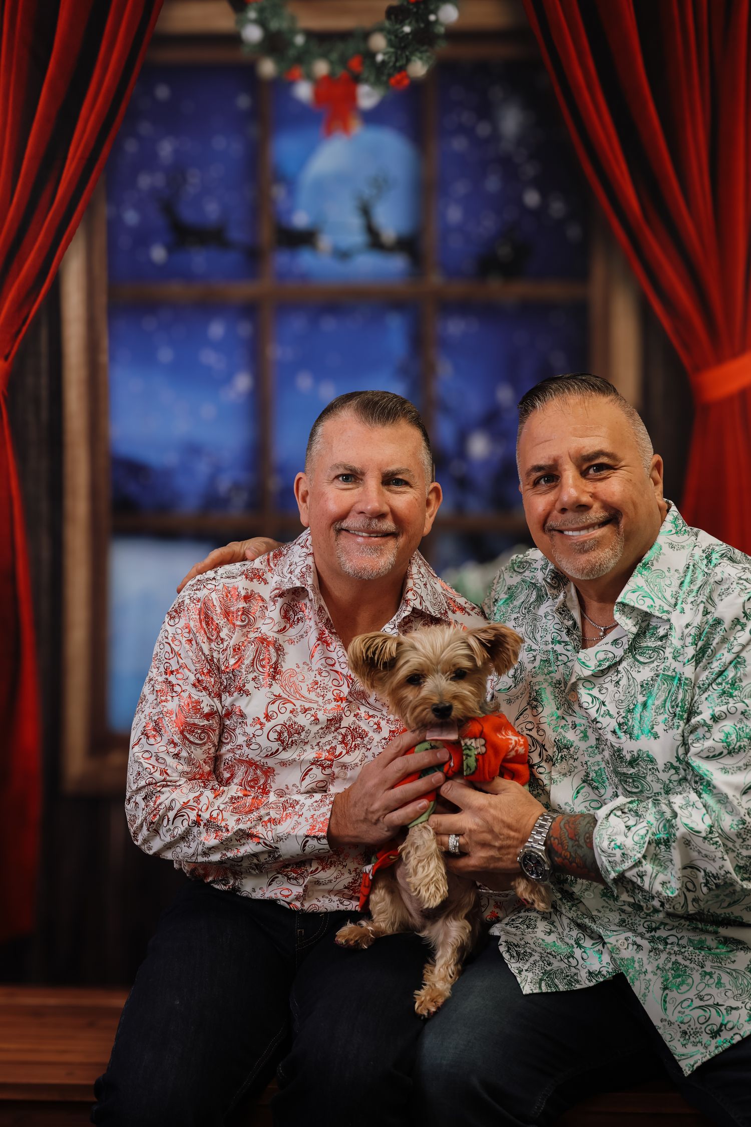 Two men in festive shirts pose with a Yorkshire Terrier in a Christmas bow at a holiday photo backdrop.