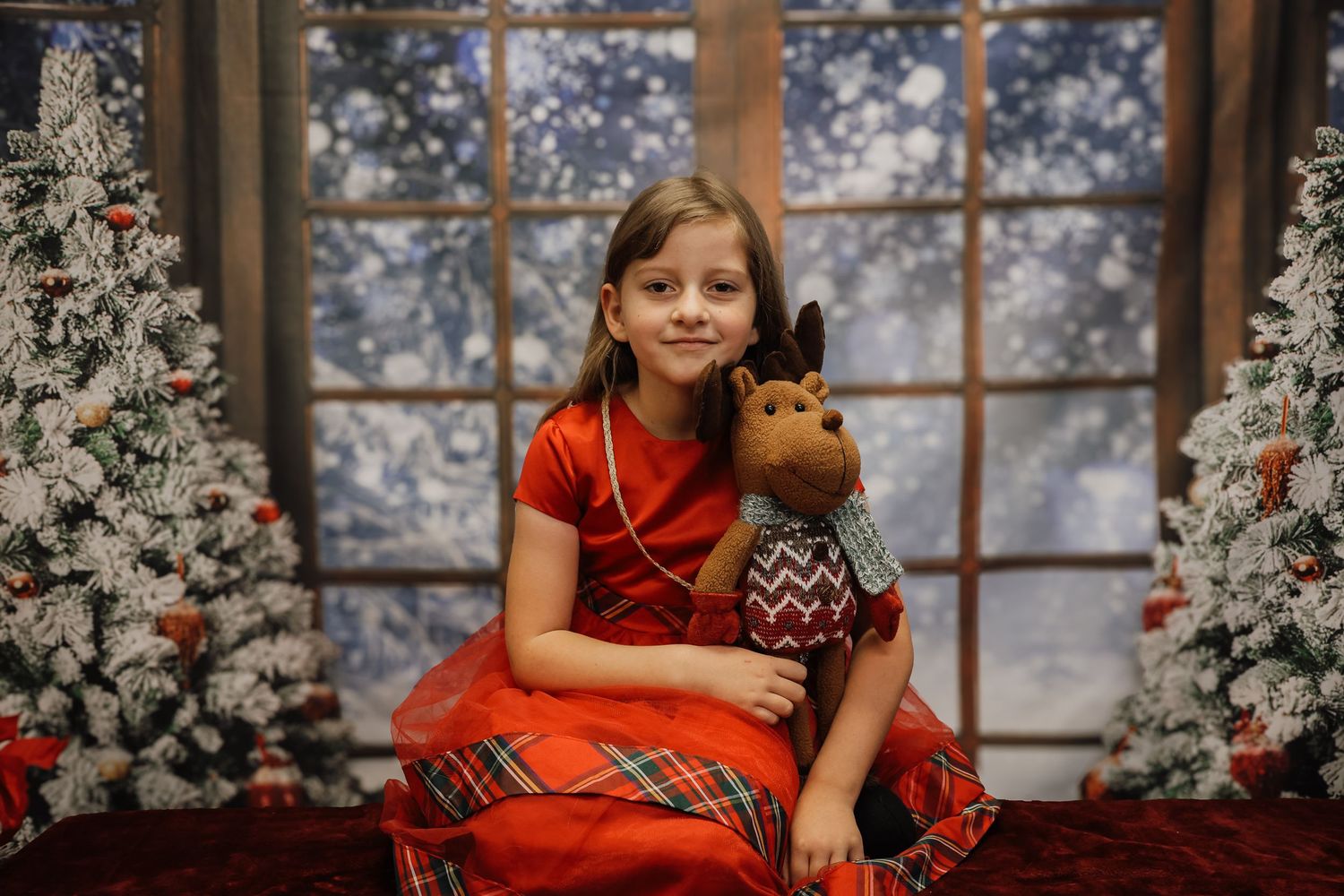 Girl in red dress holding reindeer stuffed animal poses between Christmas trees with snowy window backdrop.
